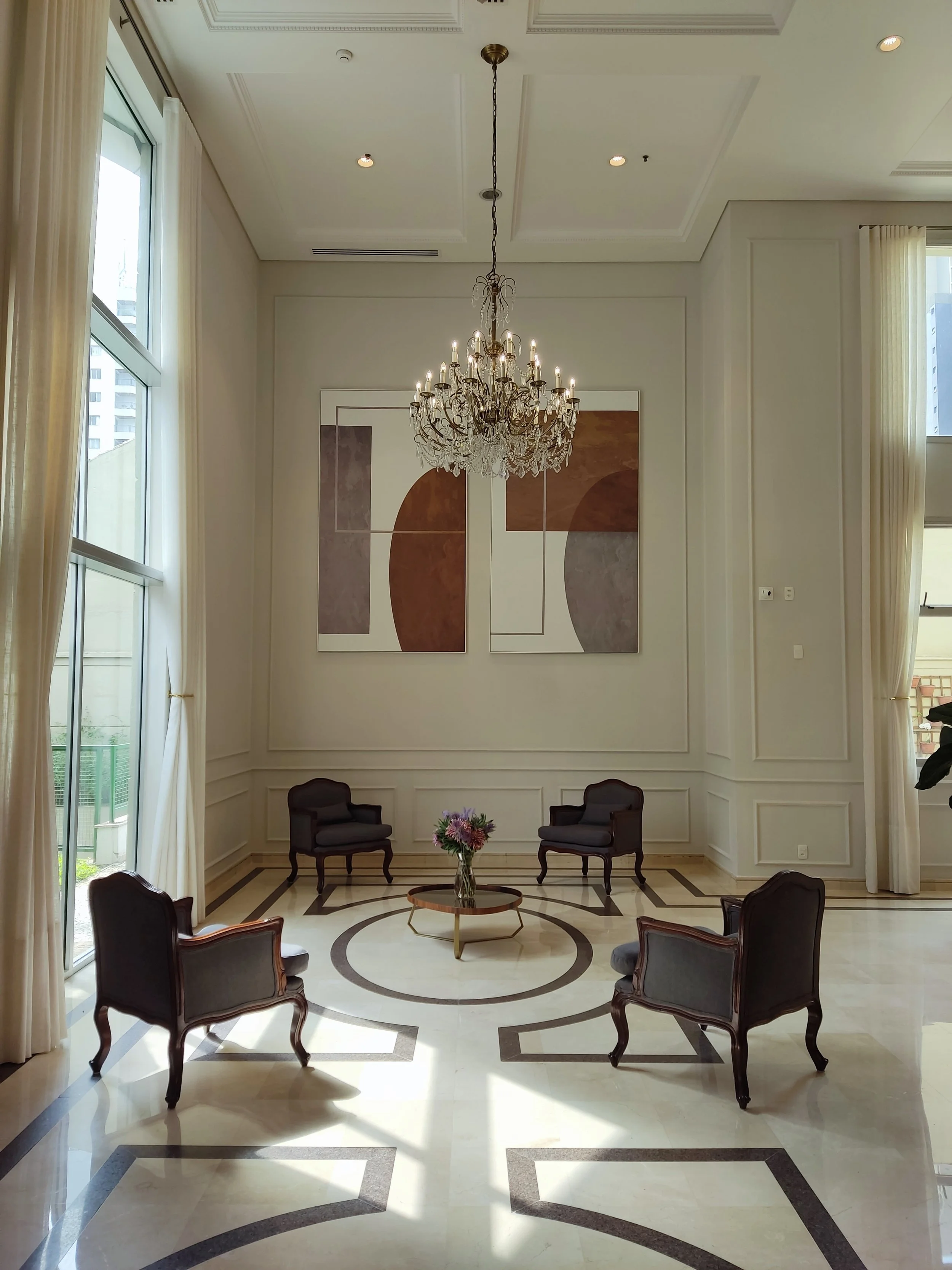 Sala de estar elegante com cinco cadeiras de madeira com estofado escuro, uma mesa redonda com um vaso de flores no centro, obras de arte abstratas na parede e um lustre de cristal pendurado no teto, com janelas grandes com cortinas claras.