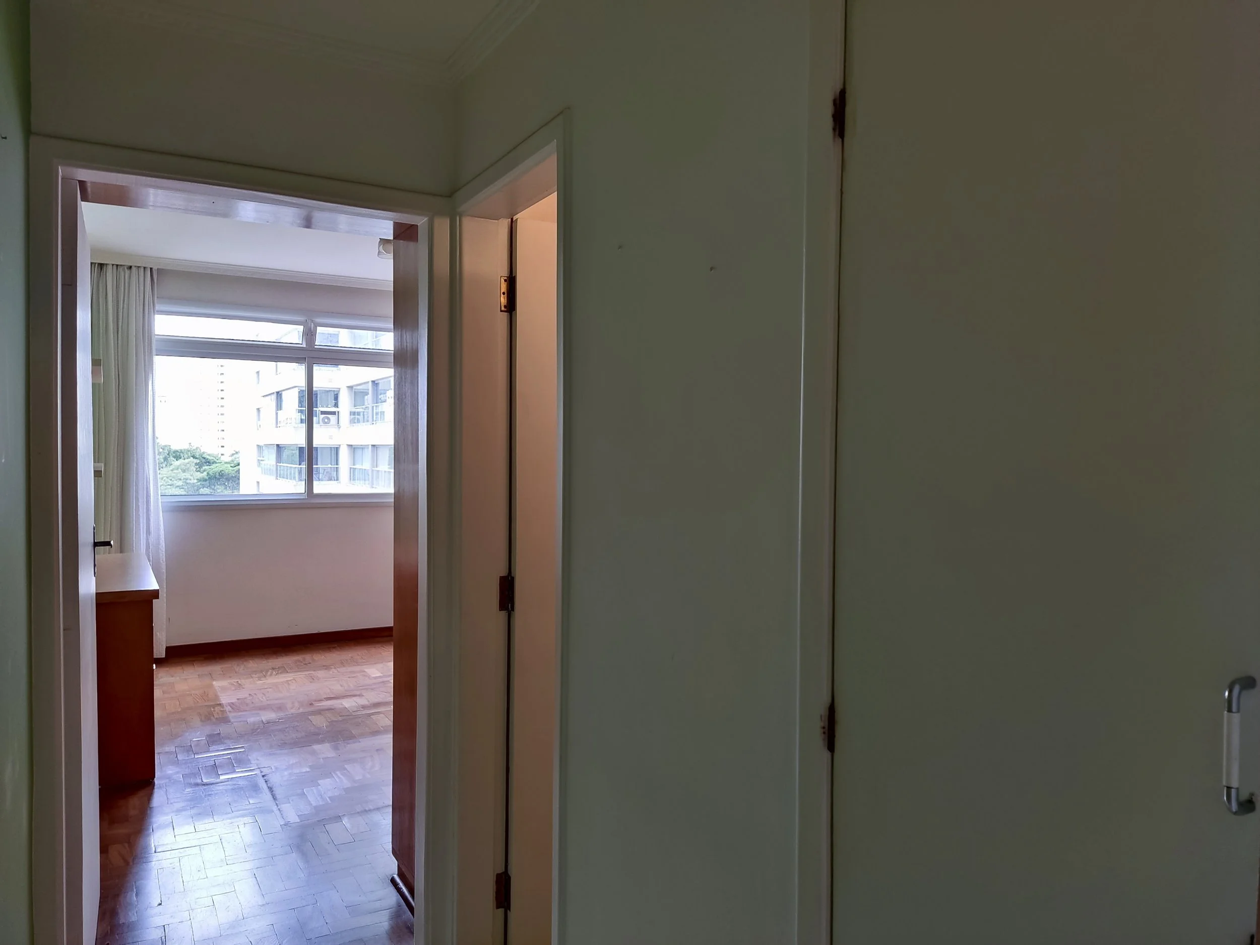 Vista de um corredor de um apartamento, com uma porta no lado direito, uma janela grande na sala ao fundo, cortinas brancas, piso de madeira e prédio visível fora da janela.