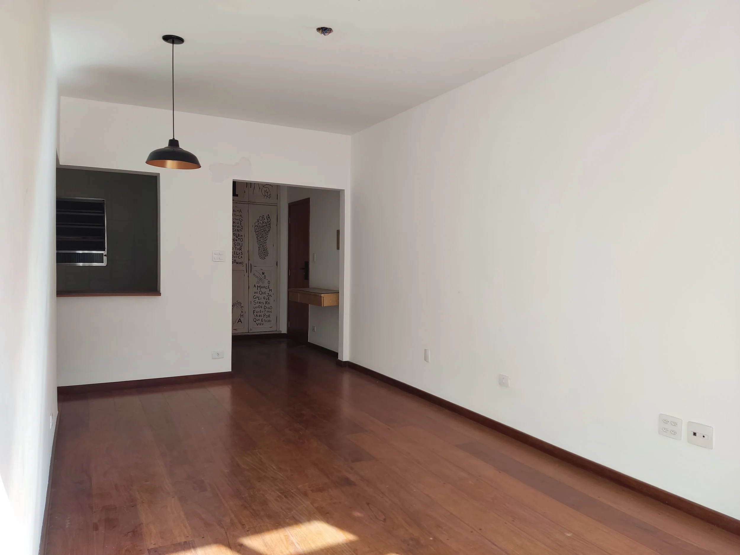 Sala vazia com paredes brancas, piso de madeira marrom e uma luminária preta pendurada no teto.