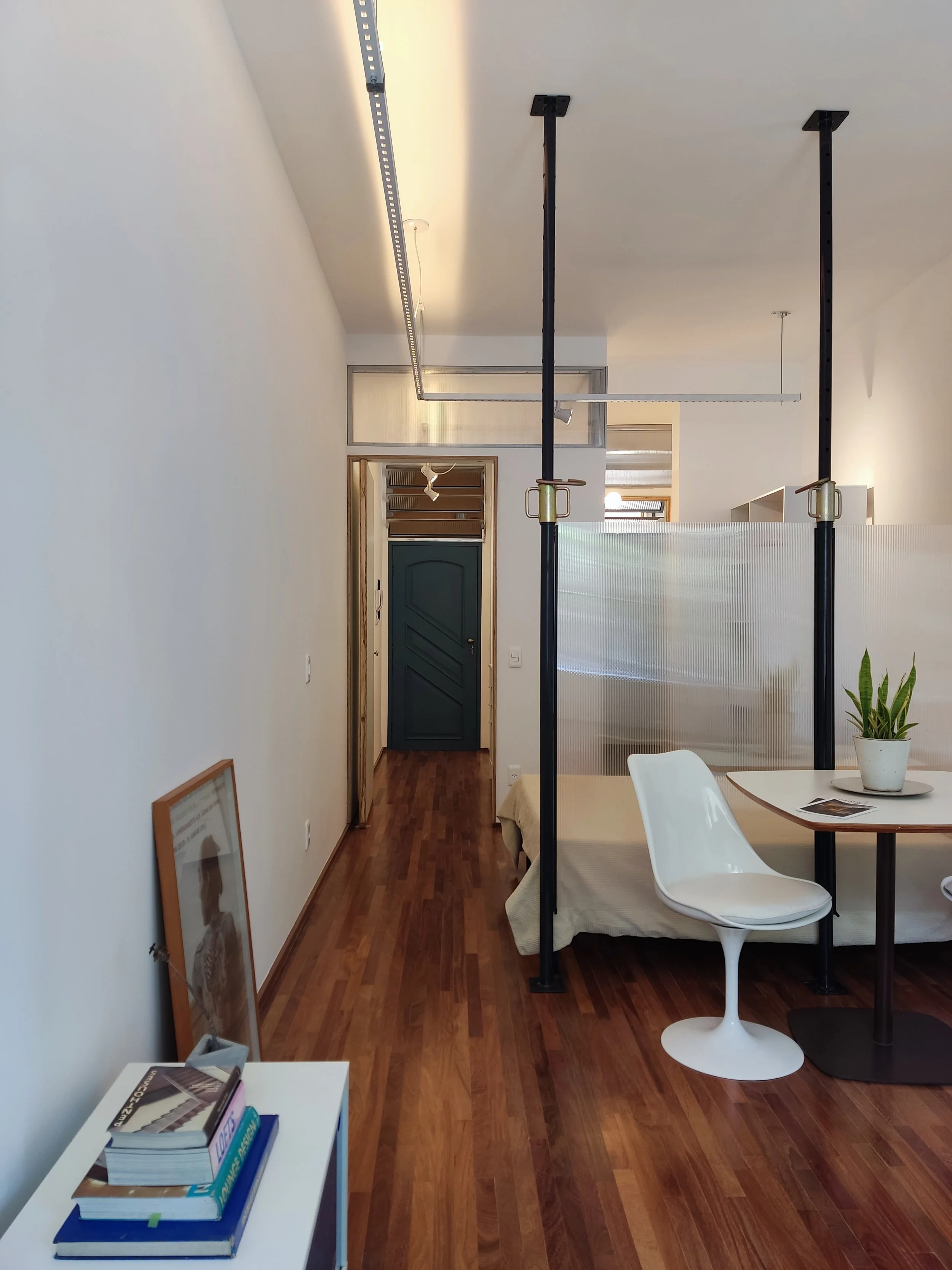 Sala de estar com piso de madeira, parede branca, mobília moderna, planta em vaso na mesa, cadeira branca de design, divisória de plástico translúcido, estrutura metálica preta para suporte, quadro apoiado no chão, livros sobre mesa