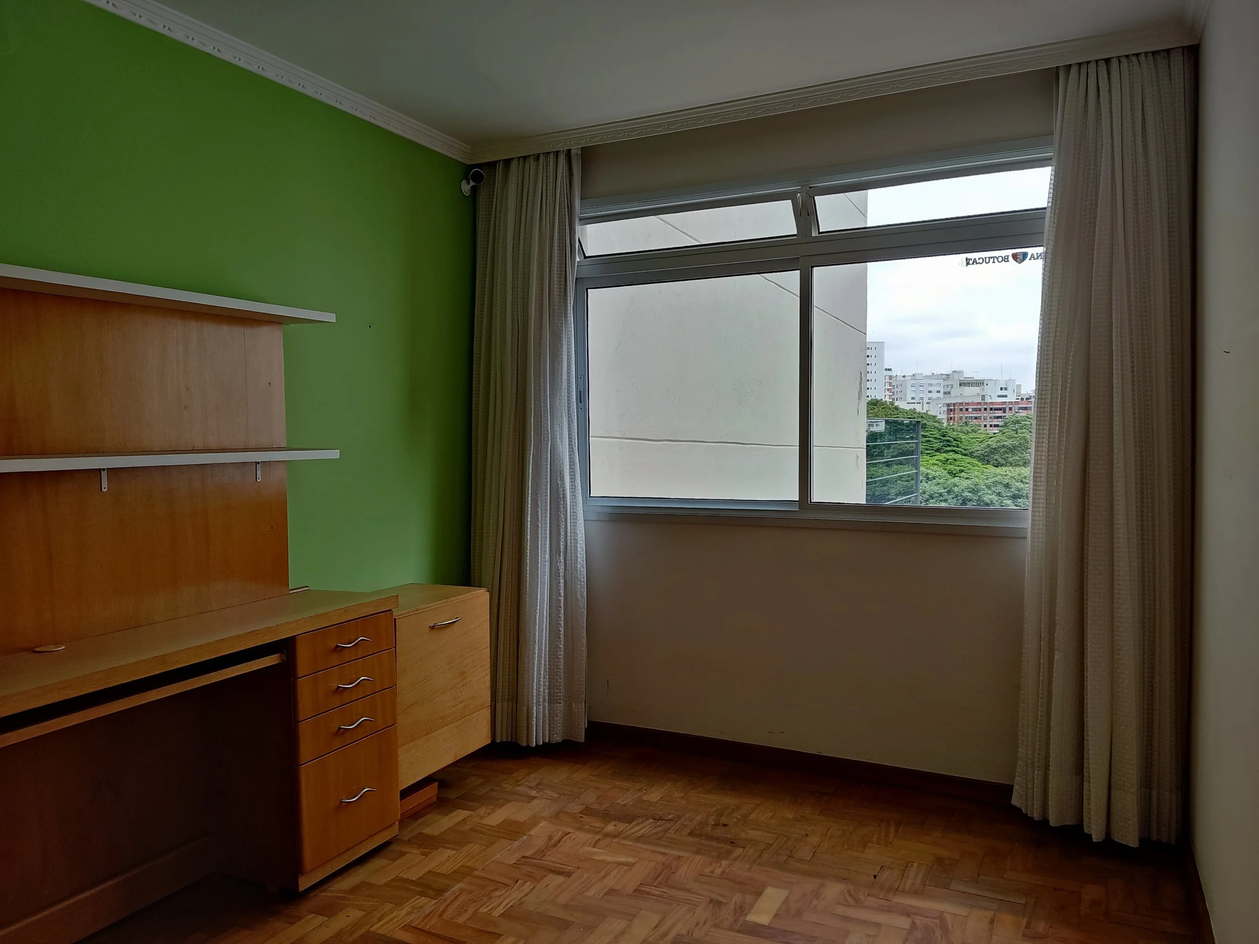 Quarto com parede verde, móvel de madeira, cortinas brancas e janela grande com vista para prédios e árvores.