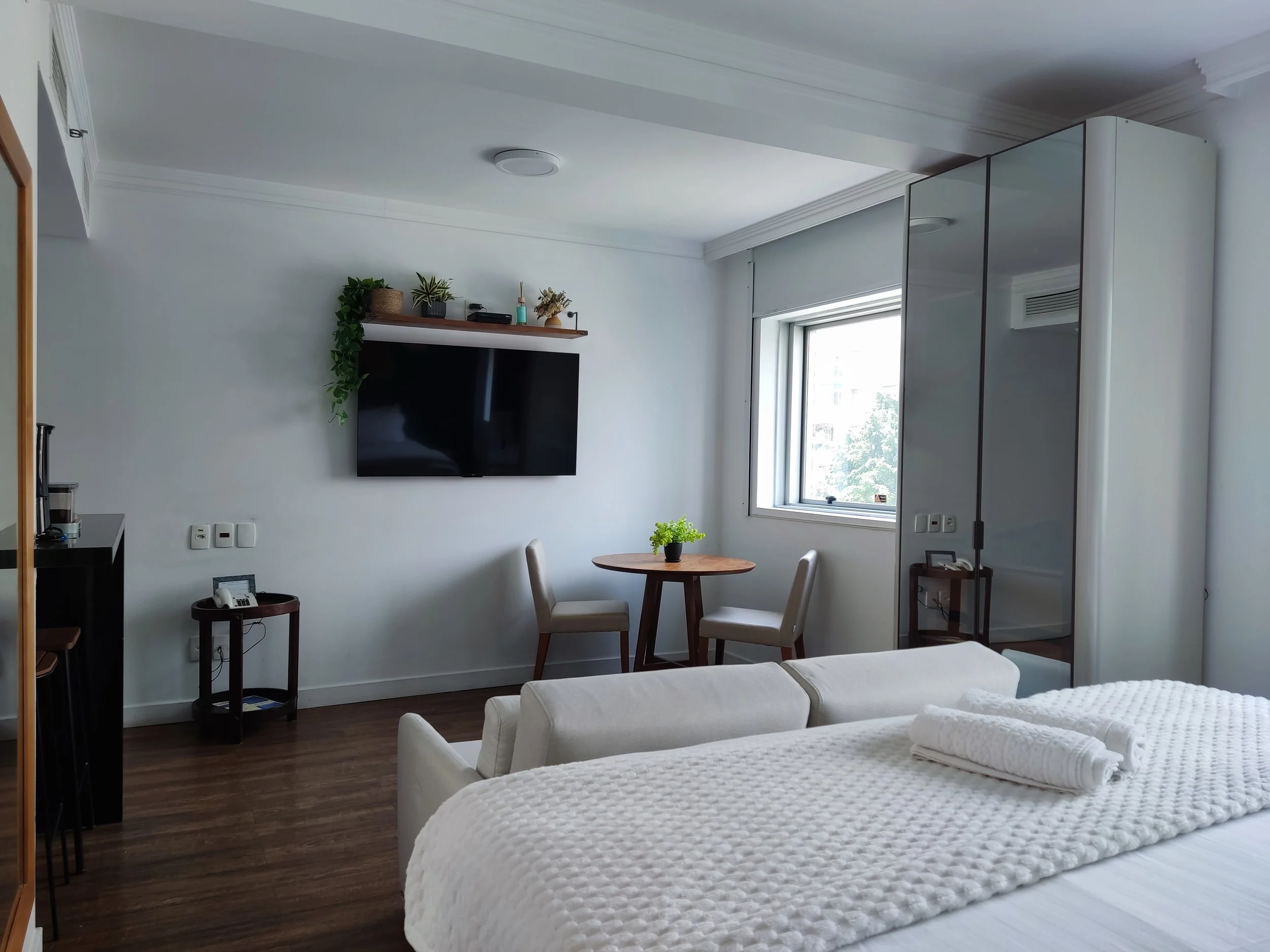 Quarto bem iluminado com cama, TV na parede, mesa redonda com duas cadeiras, armário espelhado e janela com vista para árvores.