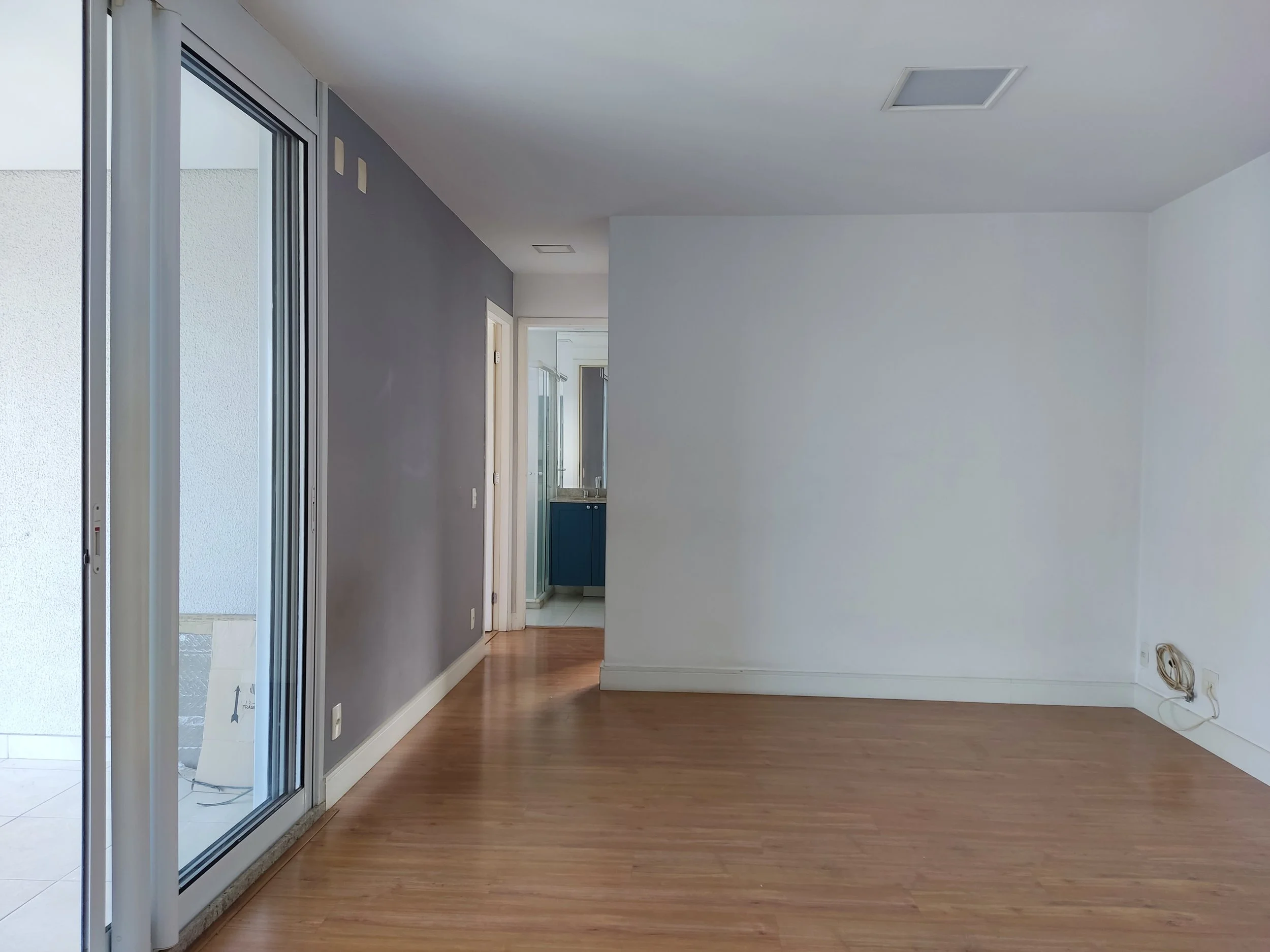 Sala vazia com piso de madeira, parede branca, parede cinza e porta de vidro que dá para uma varanda.
