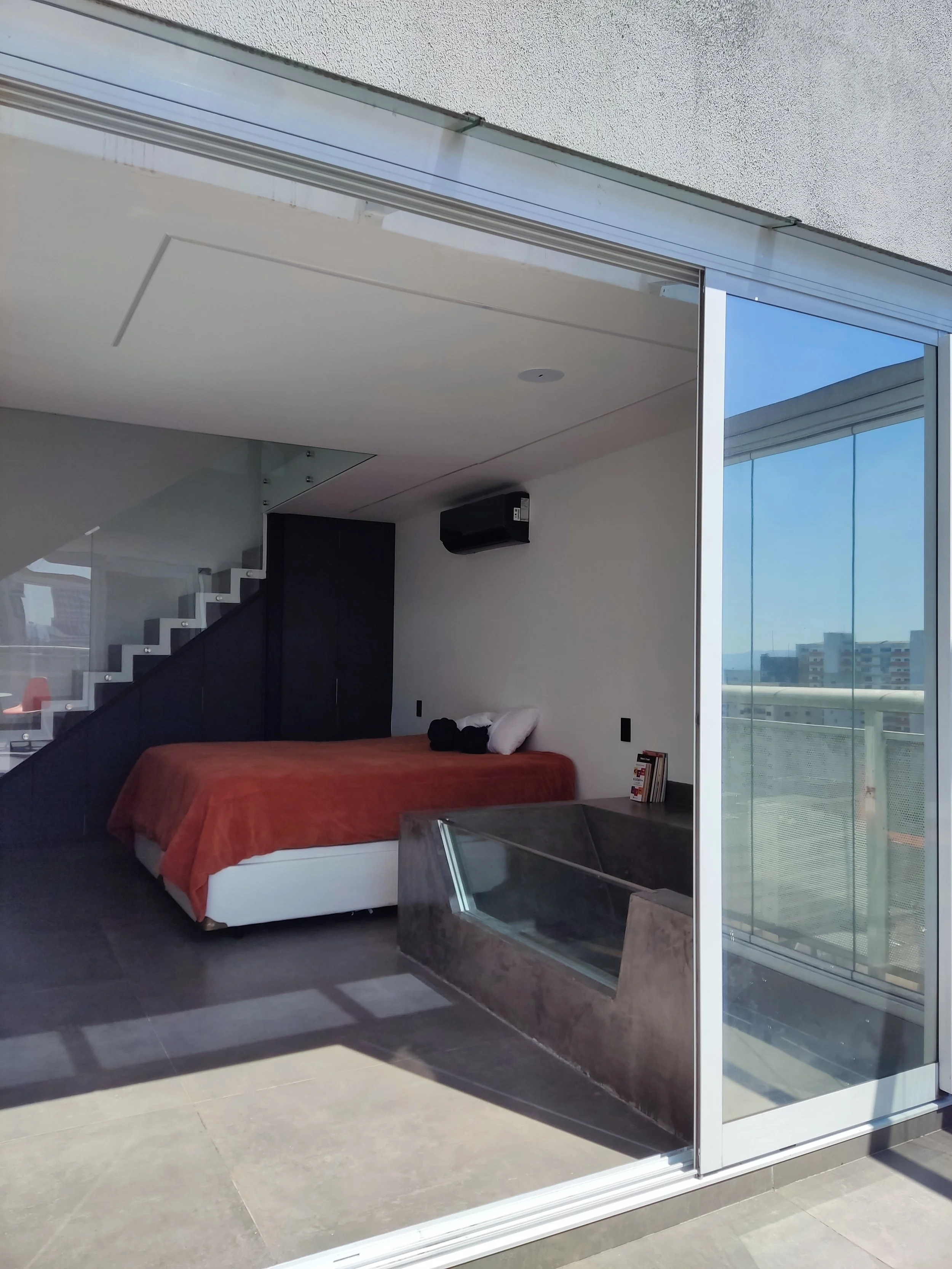 Quarto moderno com cama de casal com roupa de cama laranja, eletrodomésticos e uma escada com degraus brancos ao lado. Janela de vidro do chão ao teto com vista para edifícios e céu claro.