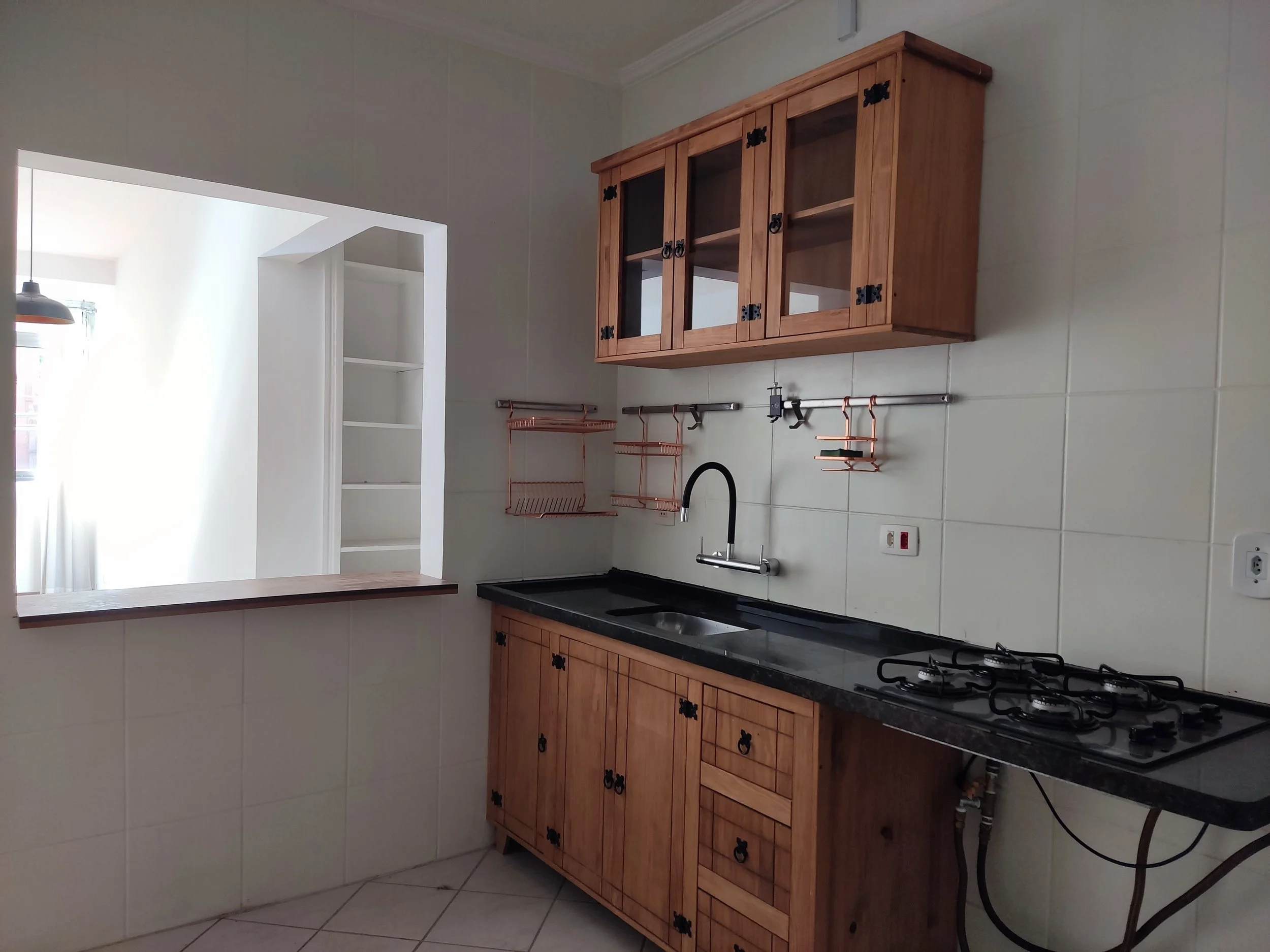 Cozinha com bancada de mármore preto, armários de madeira, teto branco, parede de azulejos brancos, uma janela de passa copos, pia com torneira preta, fogão de quatro bocas, prateleiras e armários de parede de madeira