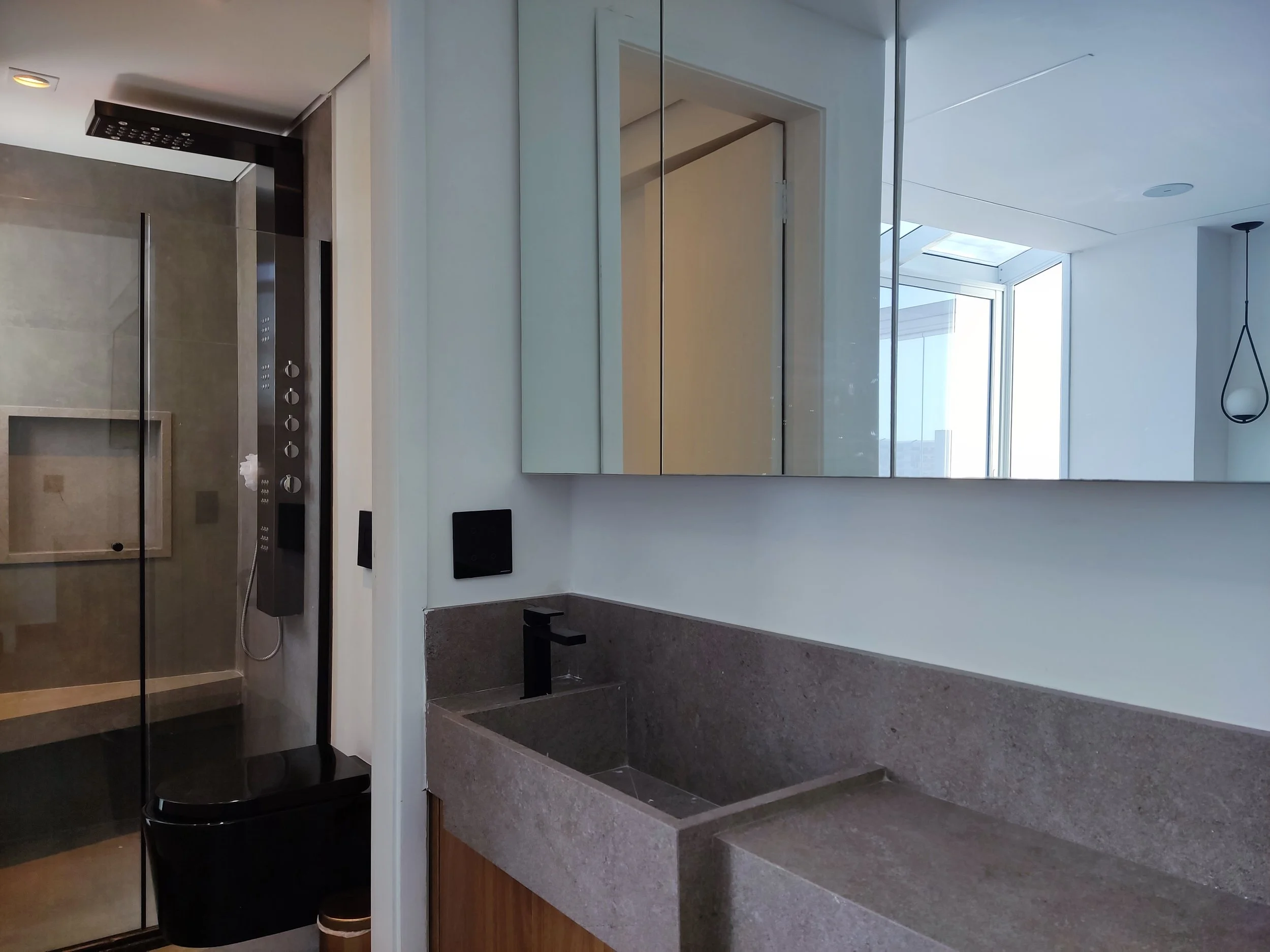 Banheiro moderno com pia de pedra, espelho grande e janela de fundo com vista para o céu e prédios, paredes brancas e iluminação natural.