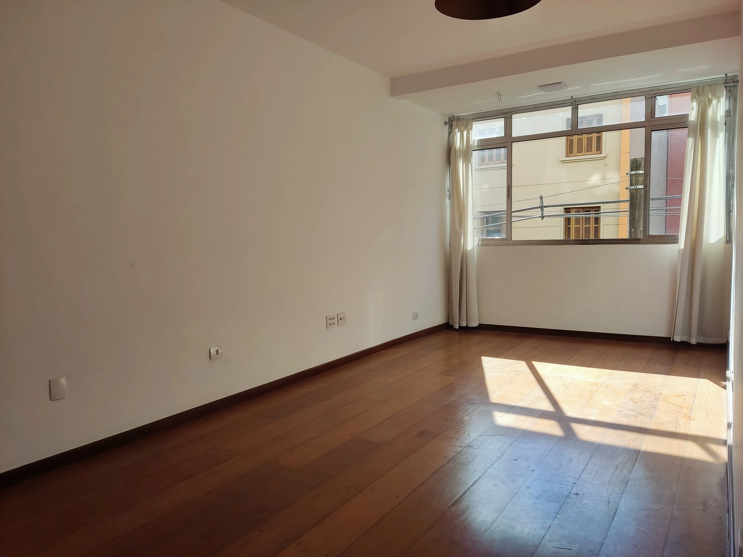Quarto vazio com piso de madeira, paredes brancas, janela grande com cortinas claras, e luz natural entrando na sala.