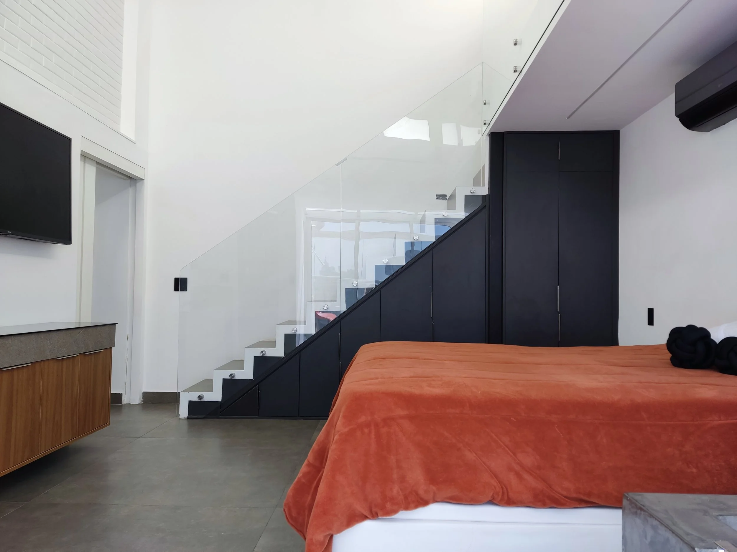 Quarto moderno com cama de baixas, roupa de cama laranja e travesseiros negros, armário escuro, televisão na parede, escada com vidro e preto, ar-condicionado e piso de cerâmica cinza.