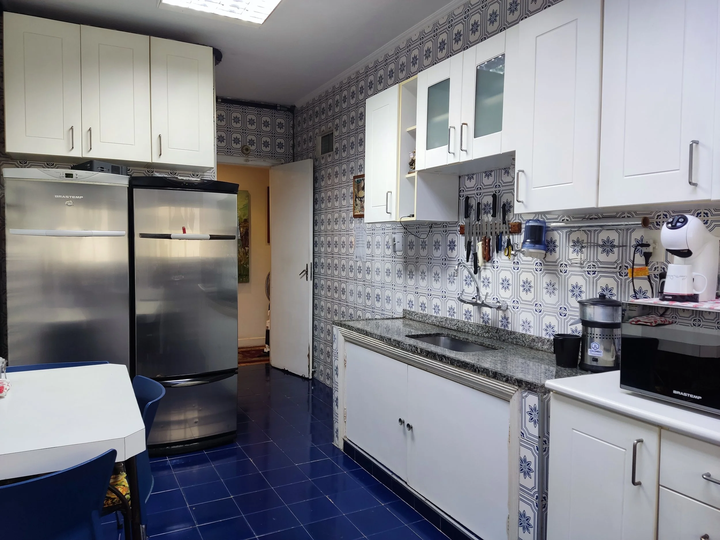 Cozinha com paredes revestidas com azulejos decorativos em azul e branco, armários brancos, geladeira de aço escovado, pia de granito, utensílios pendurados no topo da pia, eletrodomésticos como micro-ondas, cafeteira e uma garrafinha. Mesa com cadei