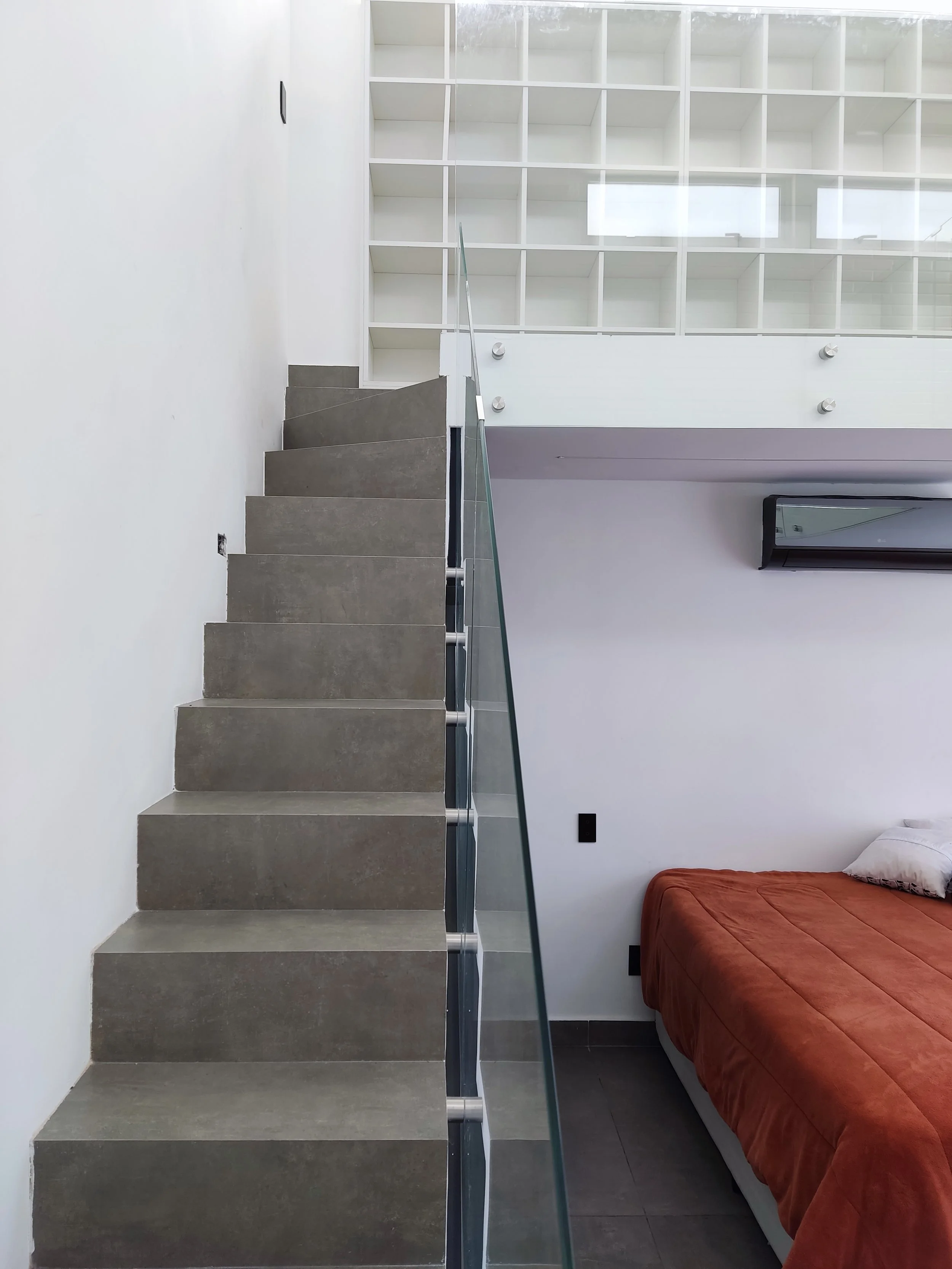 Escada de degraus com vidro de proteção ao lado de uma parede branca, em um ambiente de sala com cama de colcha em tom de tijolo e almofada branca, com parede branca e uma janela ou ar-condicionado ao fundo.
