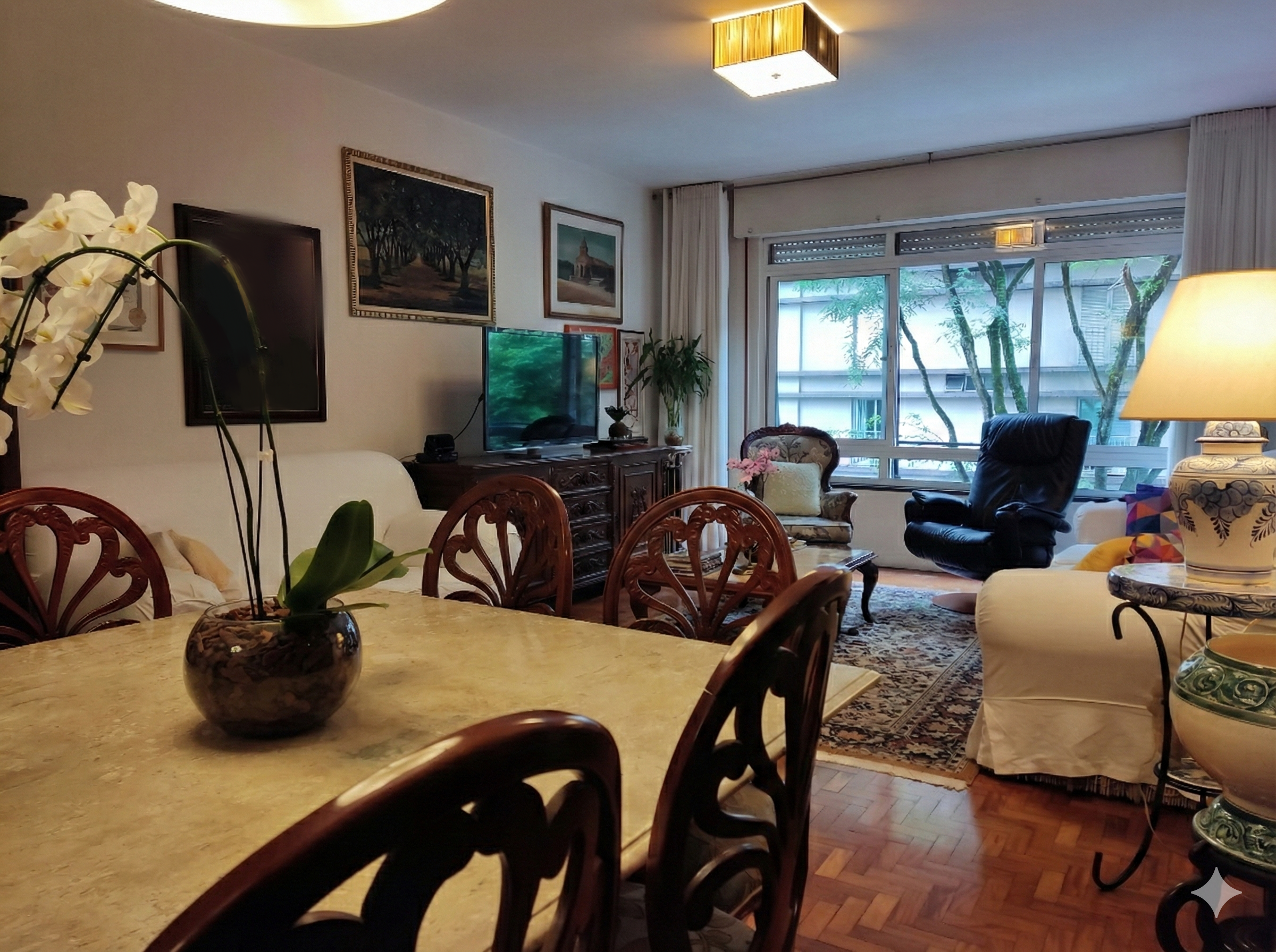 Sala de estar com mesa de jantar, sofá, cadeira de massagem, televisão, quadros nas paredes e janela grande com cortinas, decorada com plantas e luminária no teto.