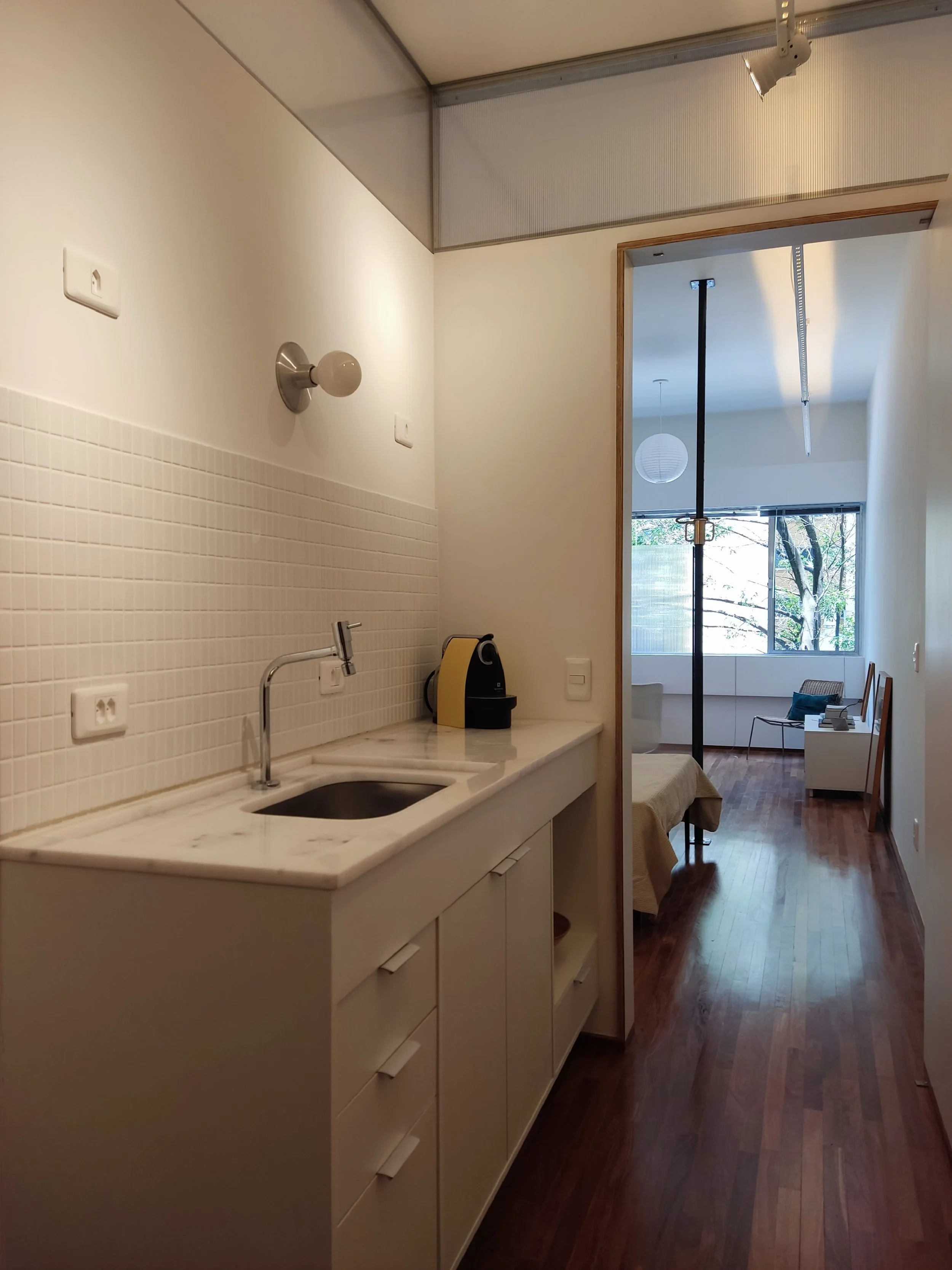 Minicozinha com pia de mármore, armários brancos e uma máquina de café na bancada. Ao fundo, sala com janelas grandes, mesa de canto e poltronas.