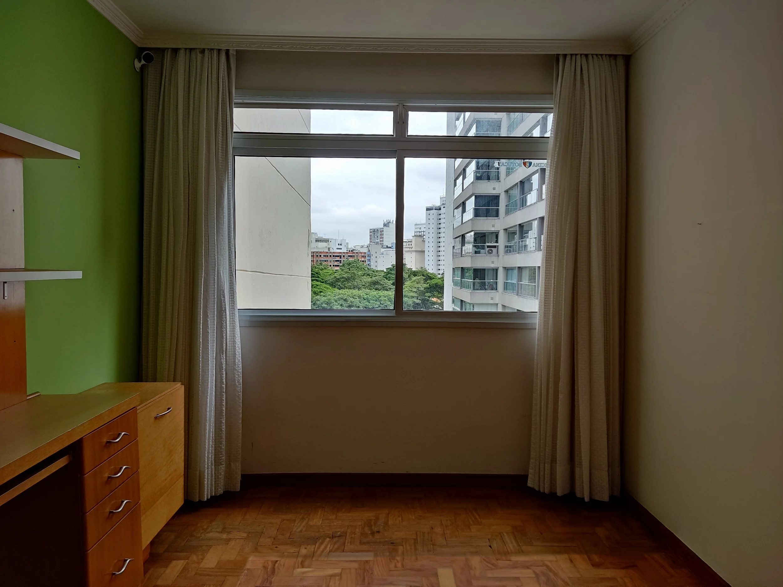Quarto vazio com janela grande, cortinas claras, parede de uma cor escura e outra clara, chão de parquet e móvel de madeira com gavetas