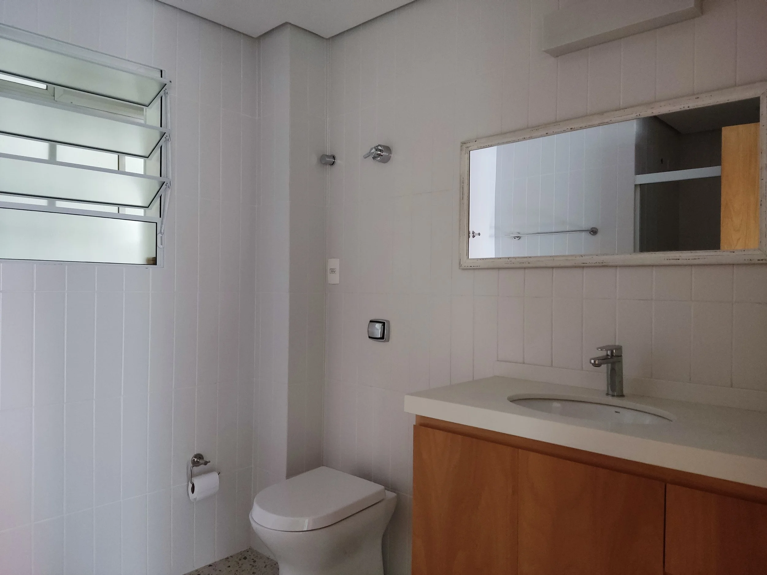 Banheiro com parede de azulejos brancos, janela de vidro com persianas, vaso sanitário, espelho grande e pia com gabinete de madeira.