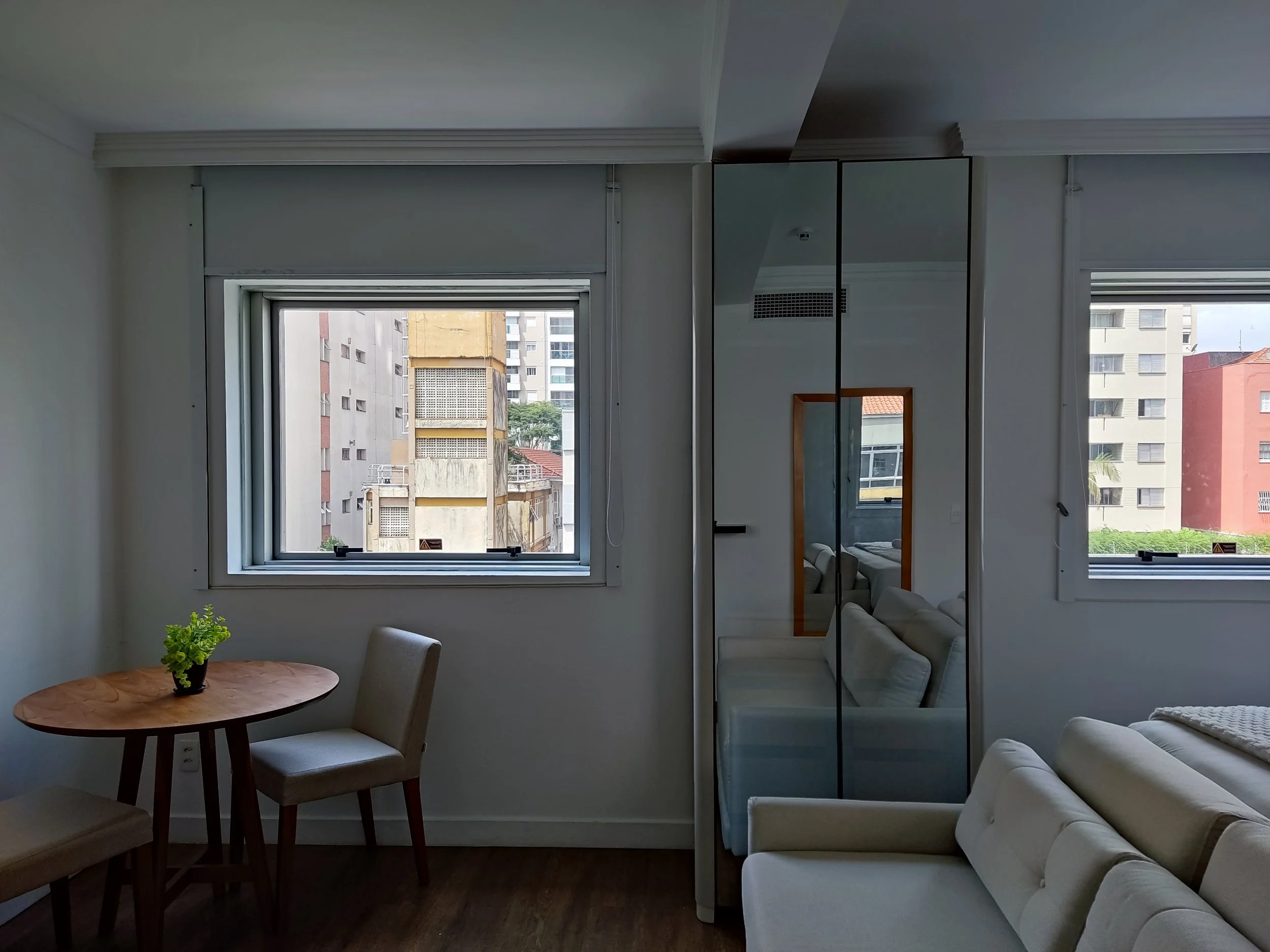 Sala de estar com janela aberta, mesa de madeira com cadeira, espelho de corpo inteiro, sofá branco e vista para prédios.
