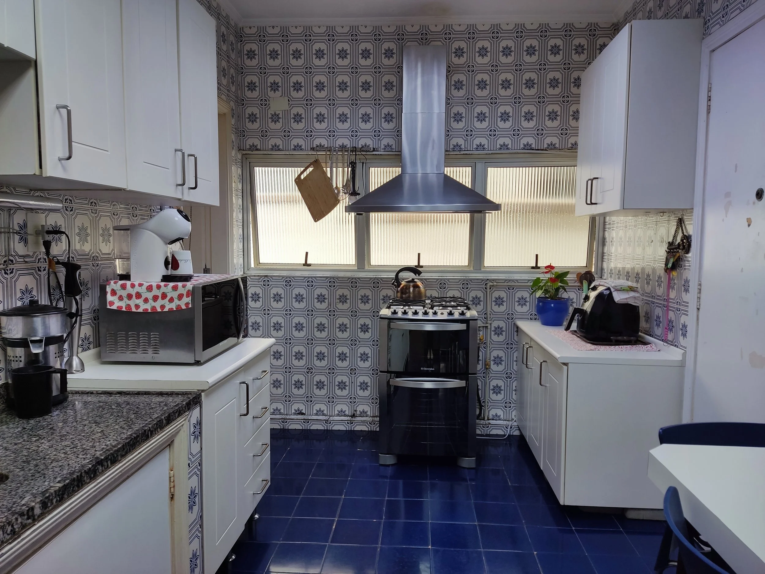 Cozinha com azulejos decorativos nas paredes, armários brancos, fogão a gás no centro, uma pia na direita, várias eletrodomésticos na bancada, uma planta colorida na bancada, janelas de vidro ao fundo, piso de azulejos azuis.