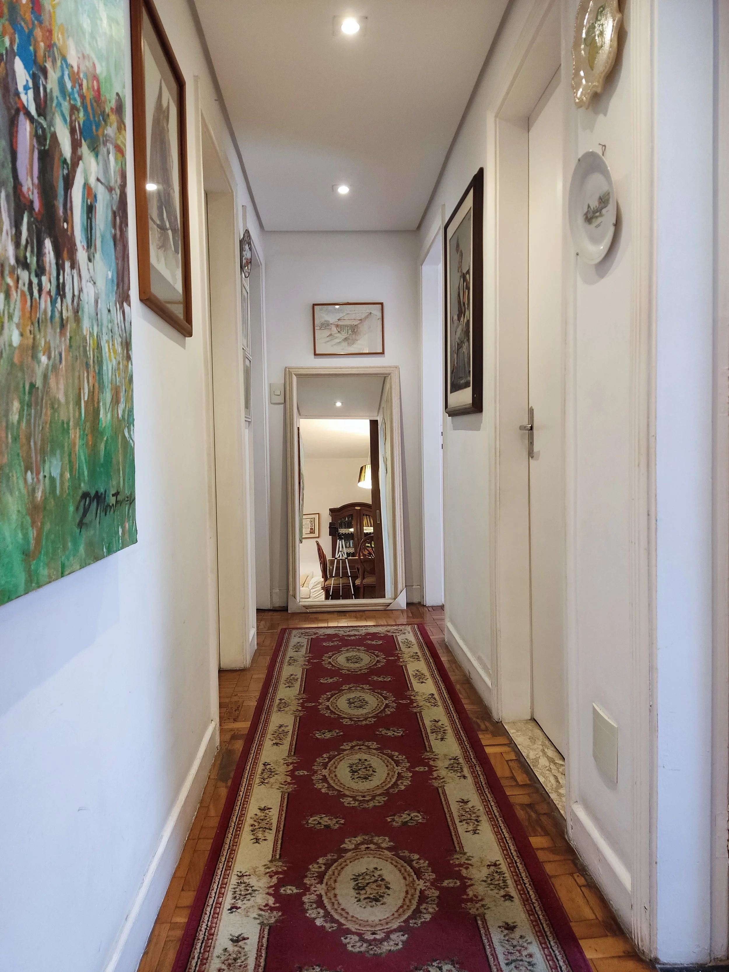 Corredor de casa com tapete vermelho com detalhes dourados e quadros nas paredes, porta de espelho na extremidade e uma porta branca à direita.