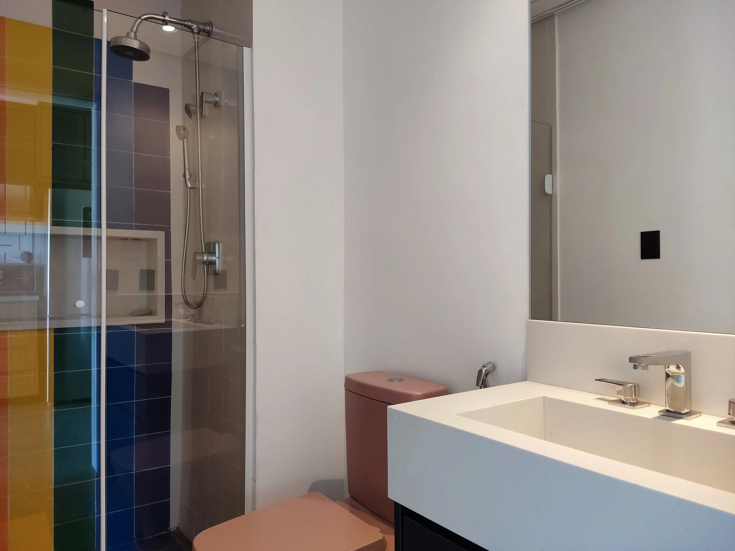 Banheiro com vaso rosa, pia branca com torneira moderna, box de vidro com porta colorida e parede branca