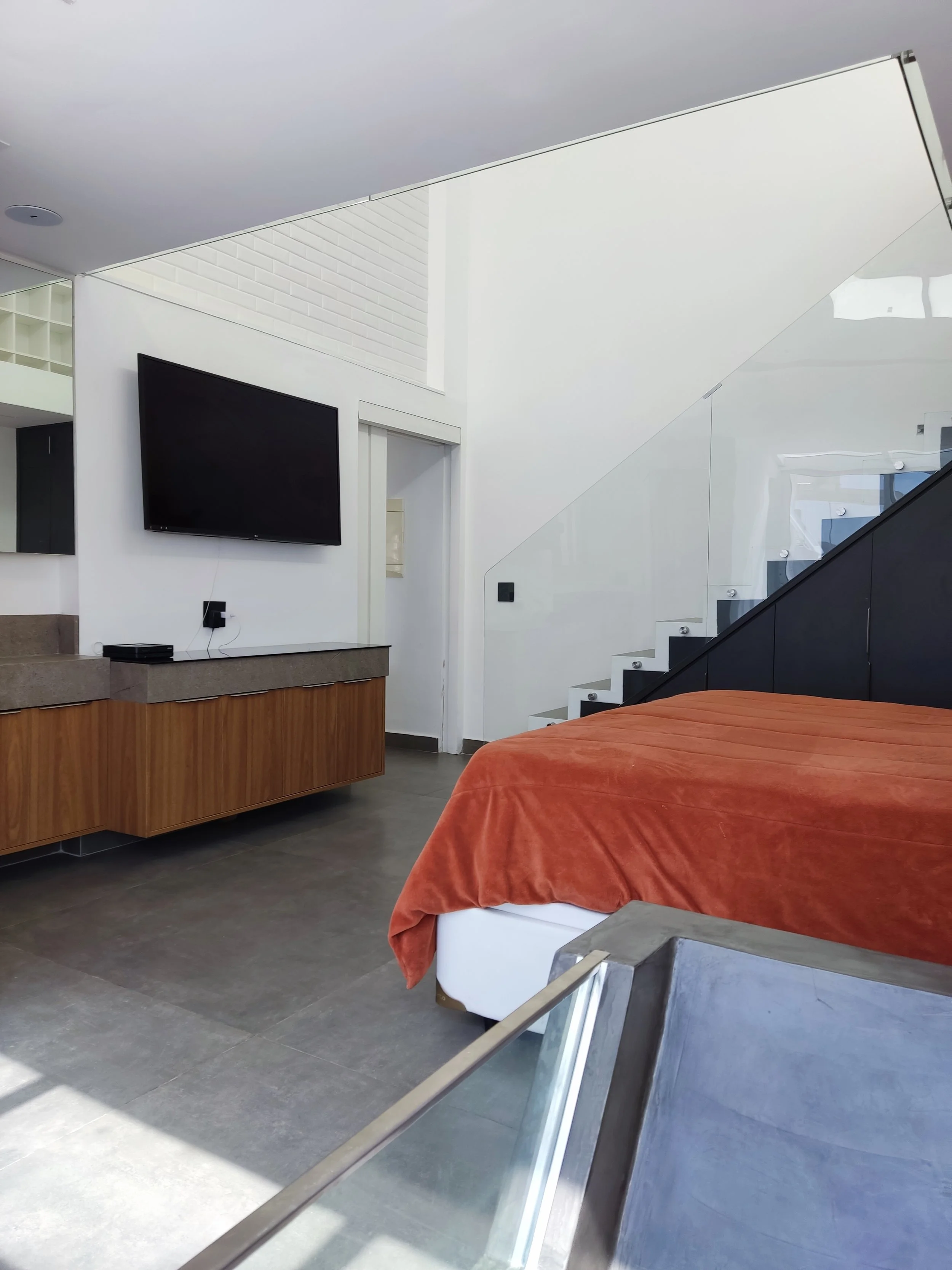 Quarto moderno com TV de tela plana na parede, cama com cobertor laranja, escada de vidro ao lado, e móveis de madeira e cimento.
