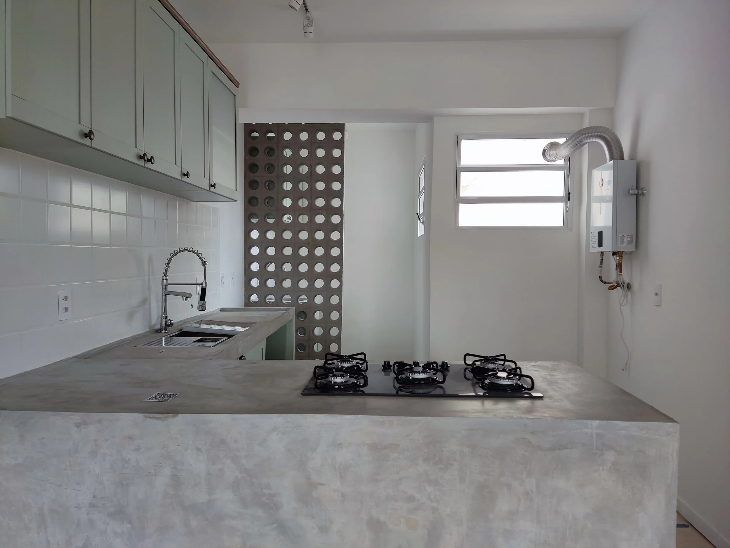 Cozinha moderna com armários verdes claros, pia com torneira de aço inox, cooktop de quatro bocas em uma bancada de concreto aparente, parede com revestimento branco e uma janela ampla com persiana, além de um aquecedor a gás na parede ao lado.