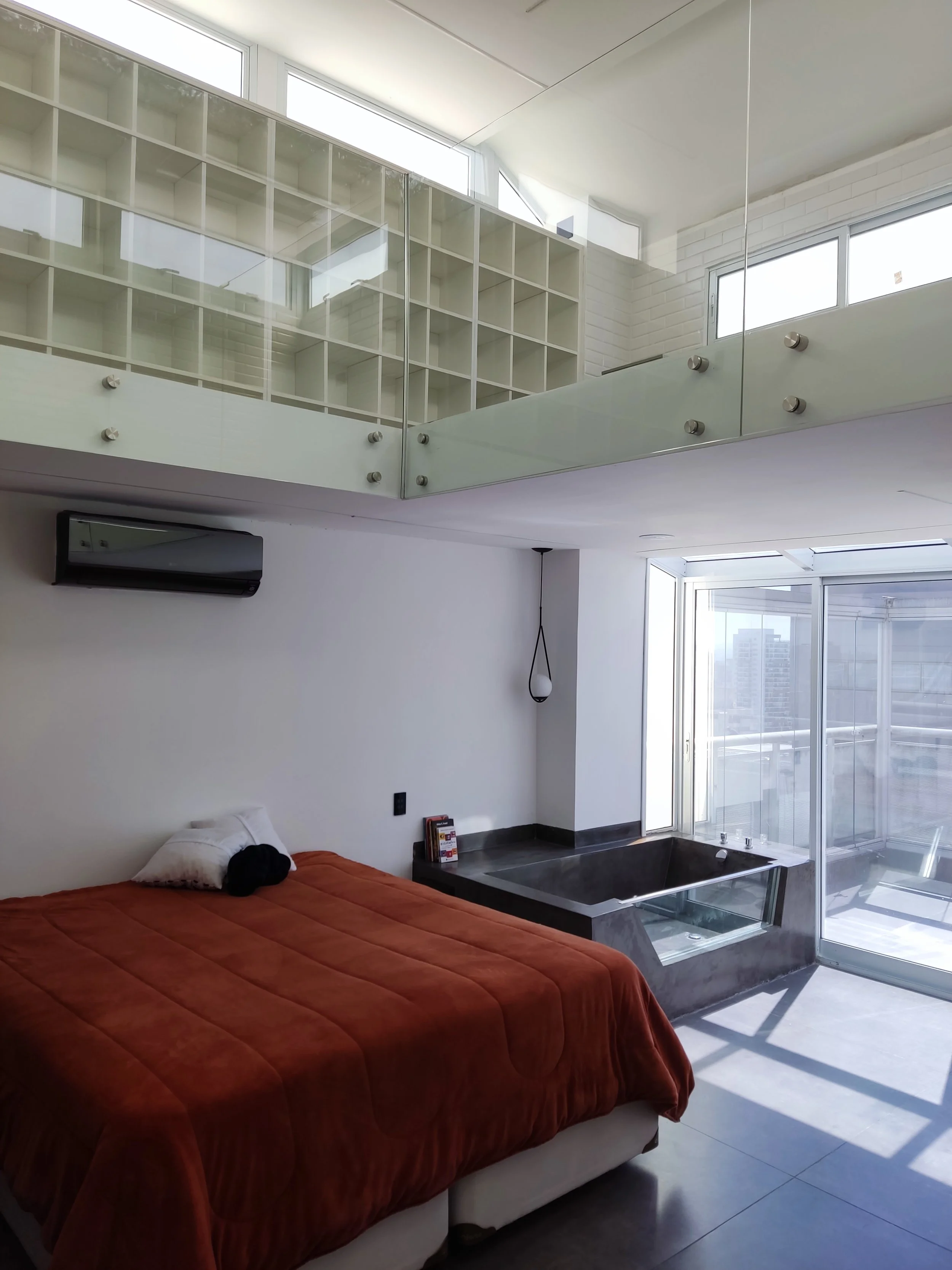 Quarto com cama de colcha laranja, janela de vidro, jacuzzi ao lado da cama, parede branca, balcão de vidro, prateleira de cubos brancos no andar de cima, iluminação natural.