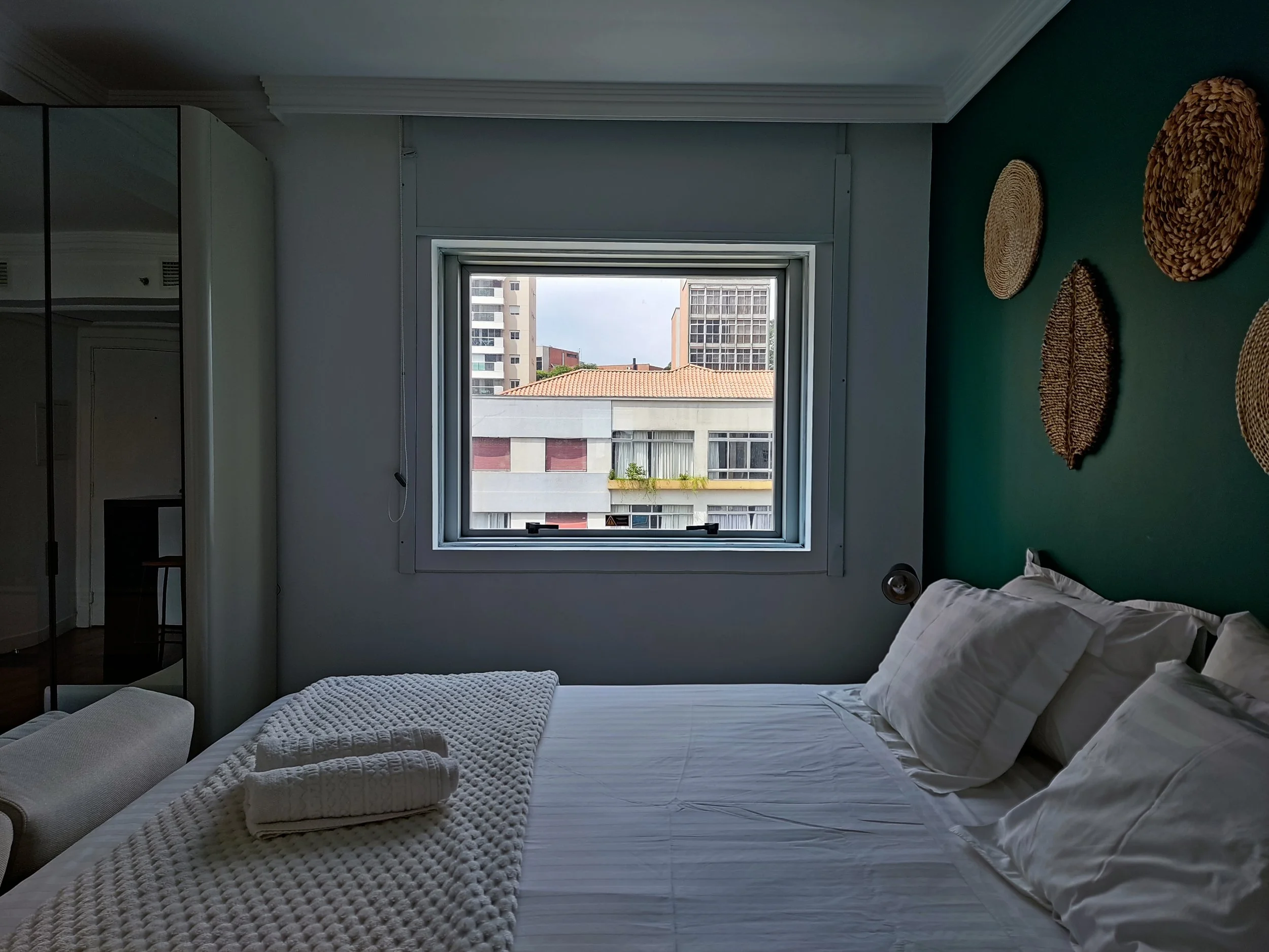 Quarto com roupa de cama branca, travesseiros e duas toalhas dobradas na cama, parede verde com objetos decorativos de fibras naturais, janela com vista para edifícios.