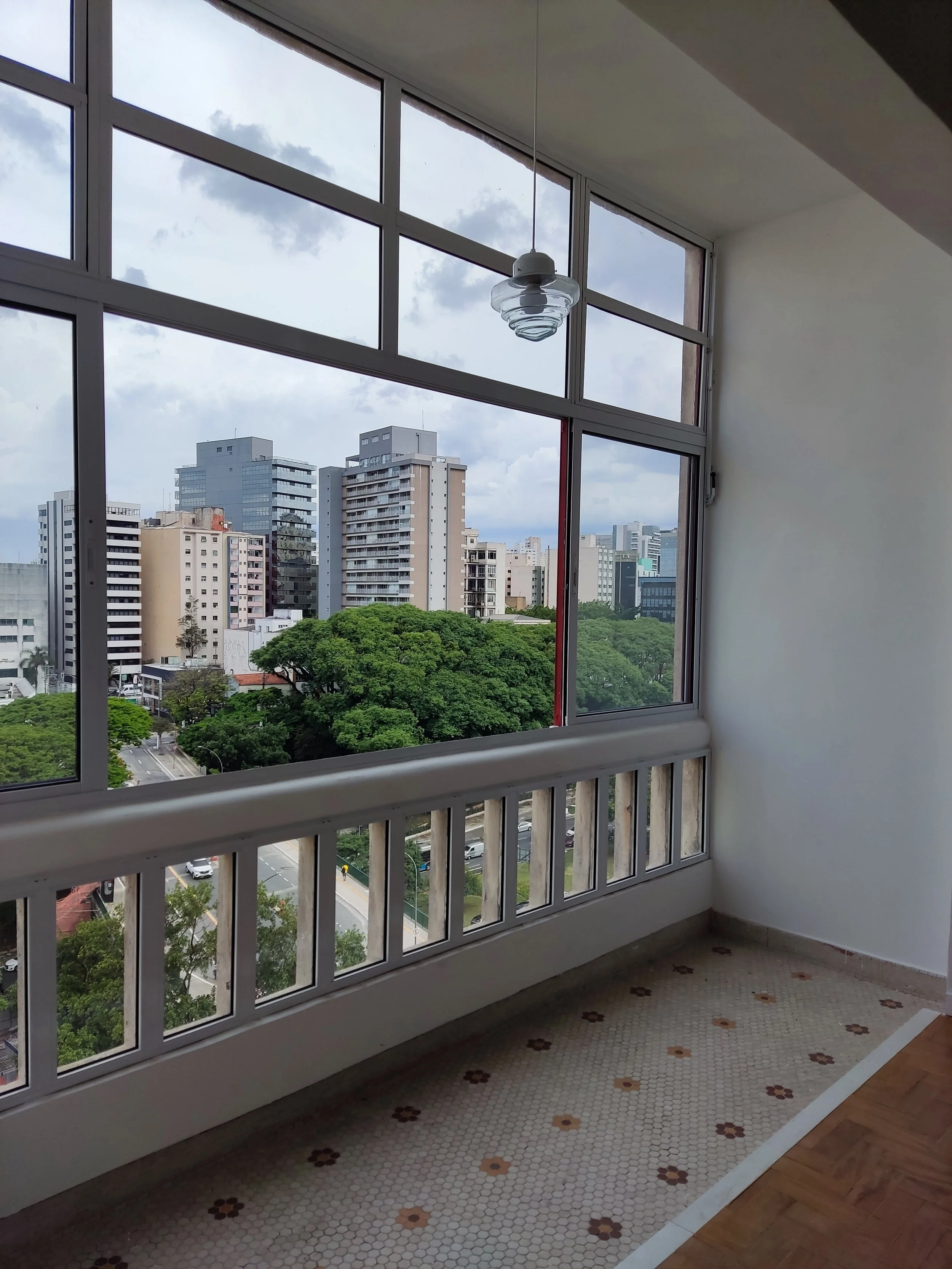 Vista de dentro de um apartamento, com janela grande de vidro, mostrando prédios altos, árvores verdes e céu nublado ao fundo.