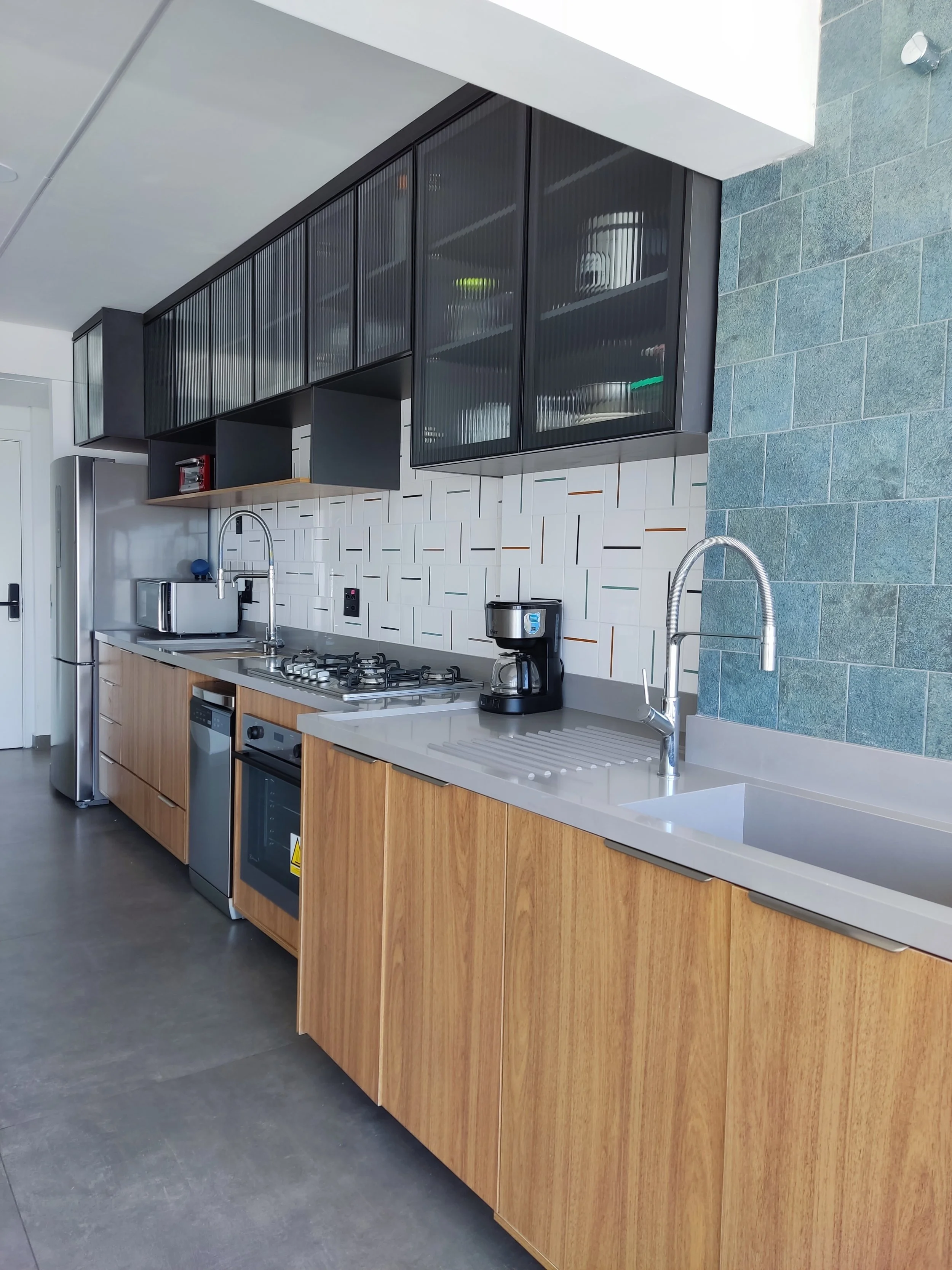 Cozinha moderna com armários de madeira, bancada cinza, backsplash de azulejos brancos e detalhes em madeira, eletrodomésticos de inox incluindo geladeira, micro-ondas, forno, cafeteira e fogão a gás, pia com torneira de aço inox, teto branco e pared