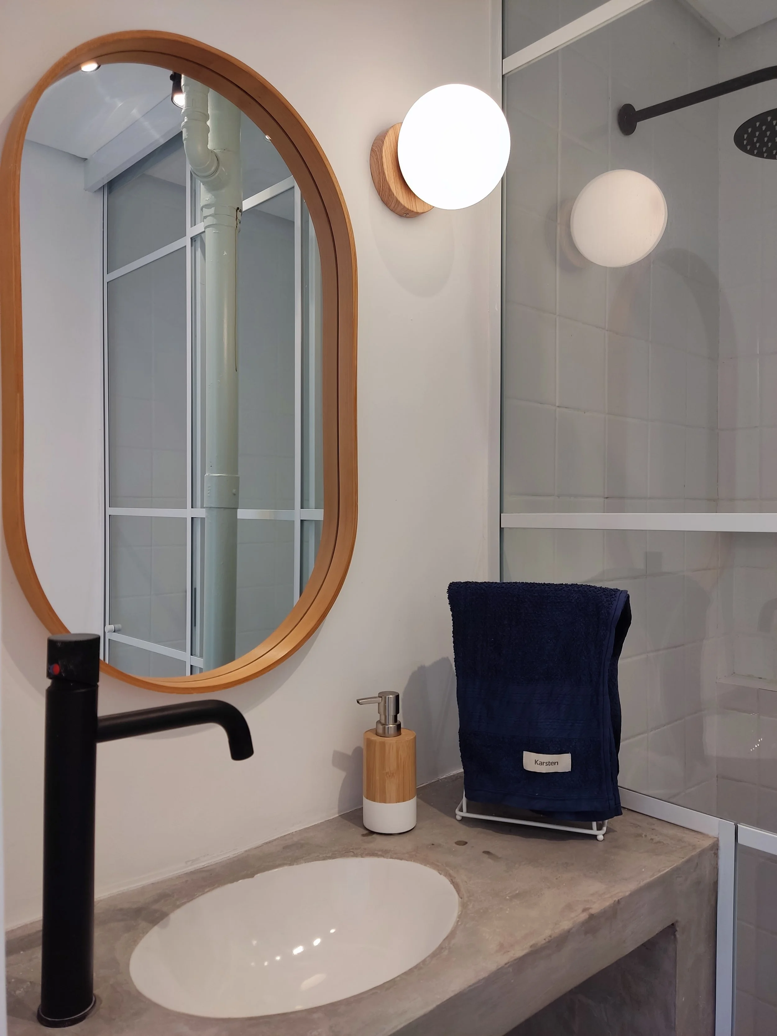 Espelho de moldura de madeira, luminária de parede de bola branca, pia de pia de banheiro com saboneteira de bambu, toalha azul escura com etiqueta Karaen, área de ducha com paredes de azulejo e vidro.