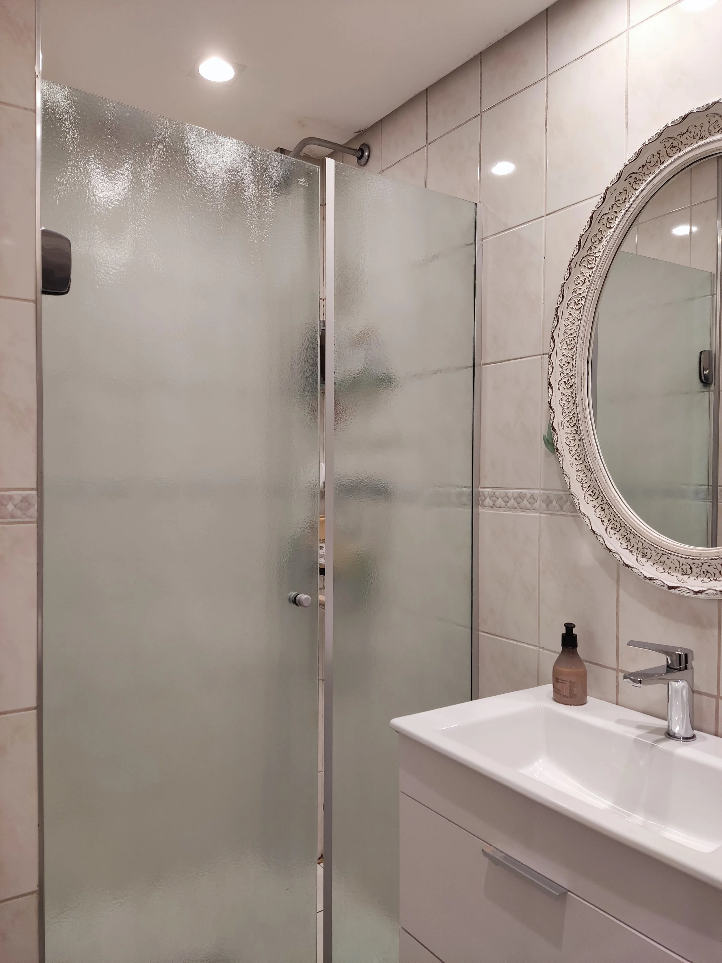 Espelho oval decorativo, pia branca com torneira, sabonete líquido no suporte, parede com azulejos claros, box de vidro fosco e teto com iluminação embutida.