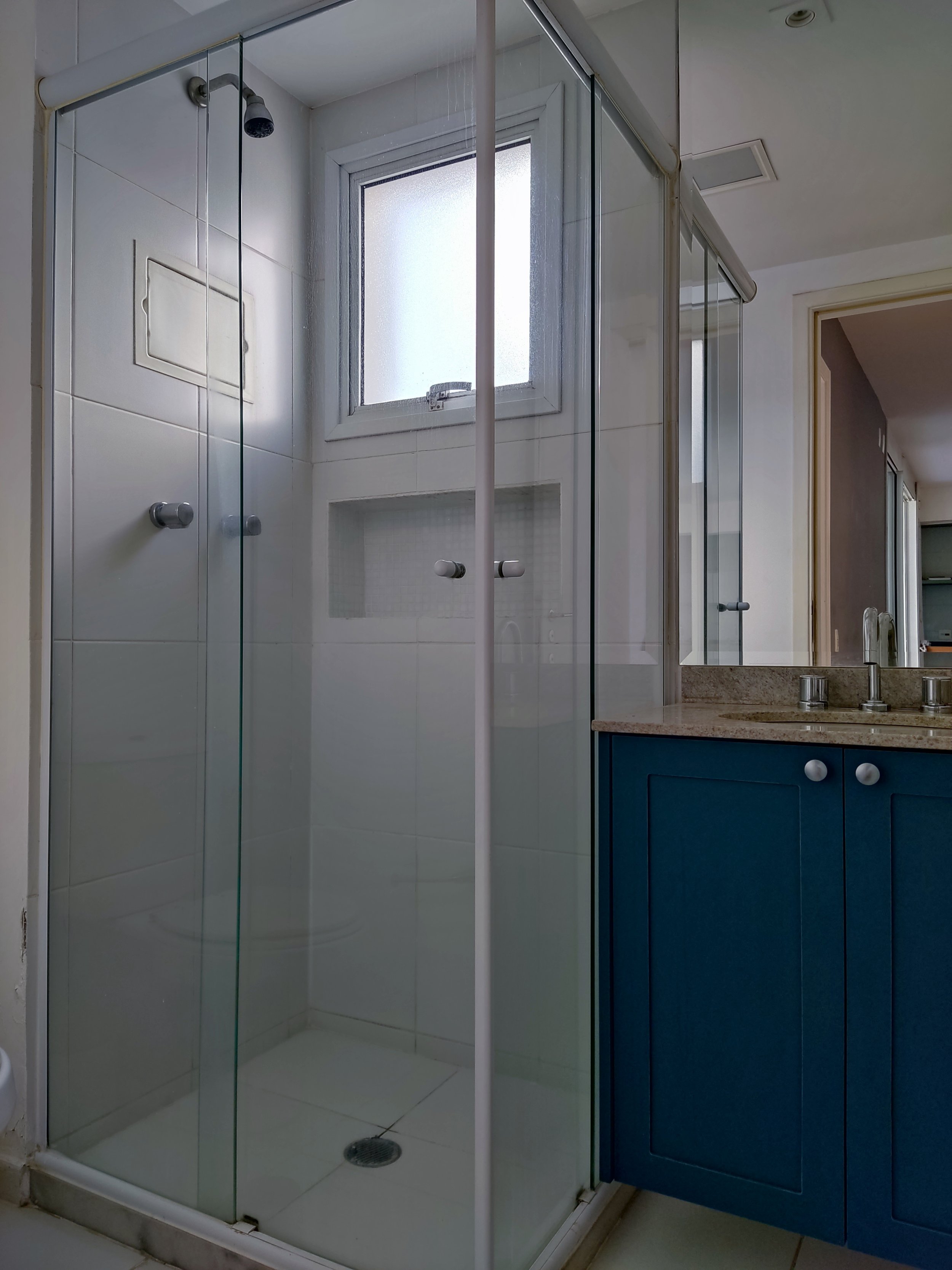 Box de vidro de chuveiro com janela de vidro ao fundo e pia com armário azul ao lado.