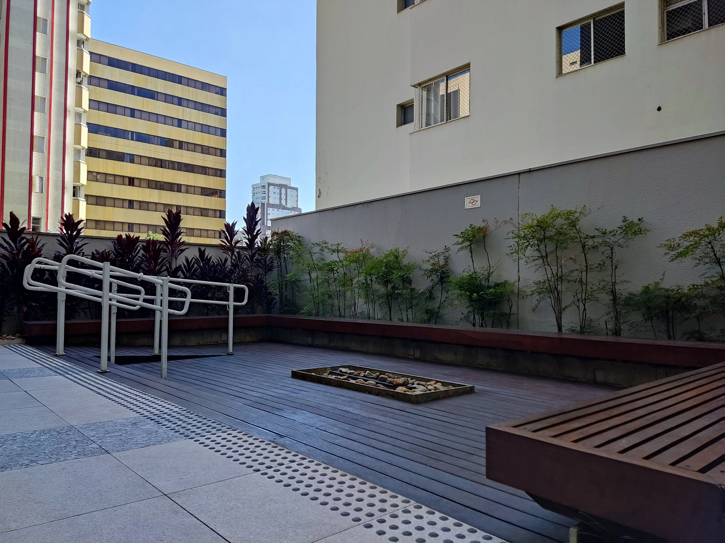Área externa de um prédio urbano com piso de cerâmica e madeira, plantas ao longo de um muro cinza, e edifícios altos ao fundo sob céu claro.