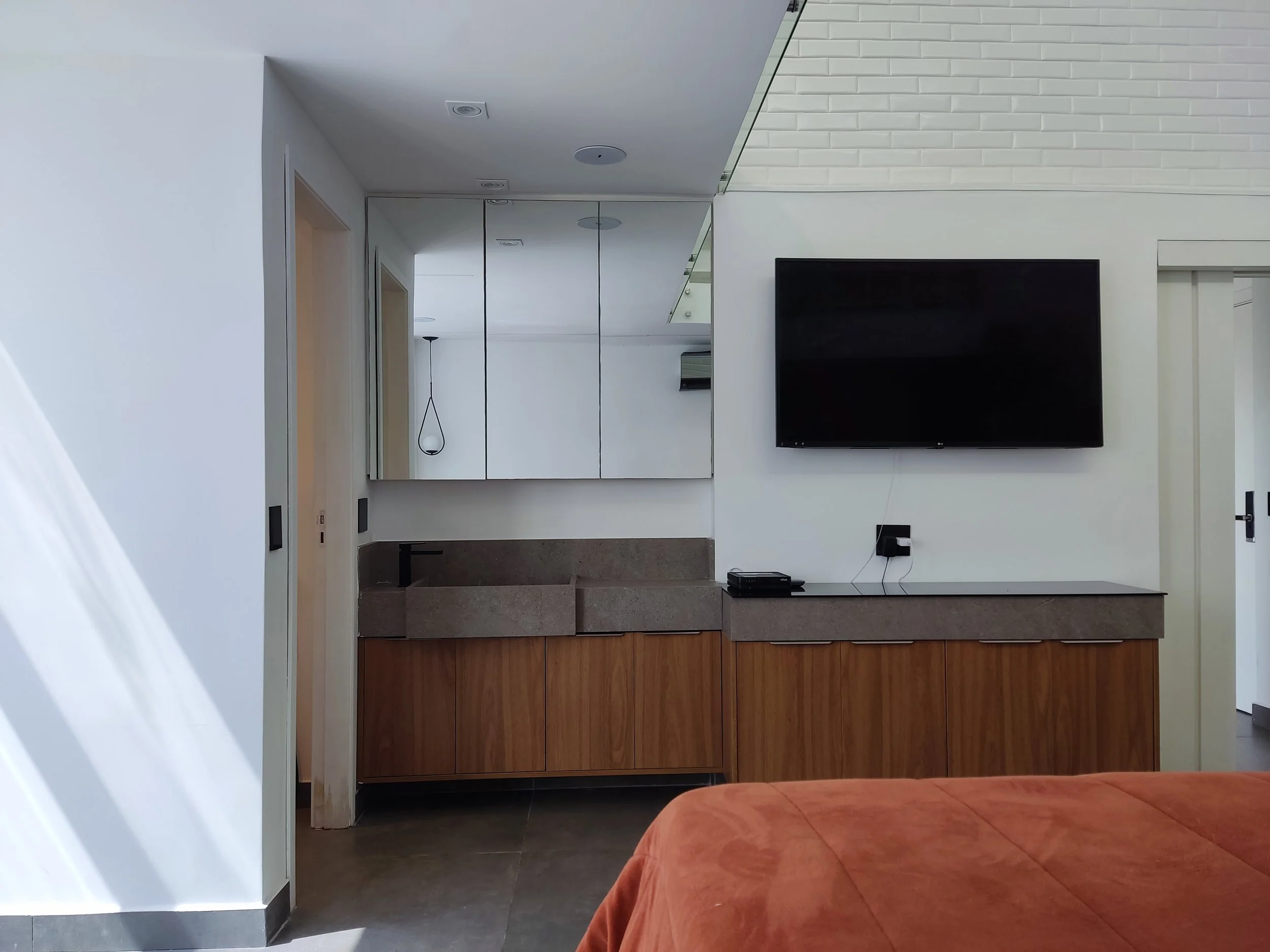 Sala de estar moderna com televisão de tela plana na parede, armário de madeira, espelho, luz pendente e colchão com capa alaranjada.