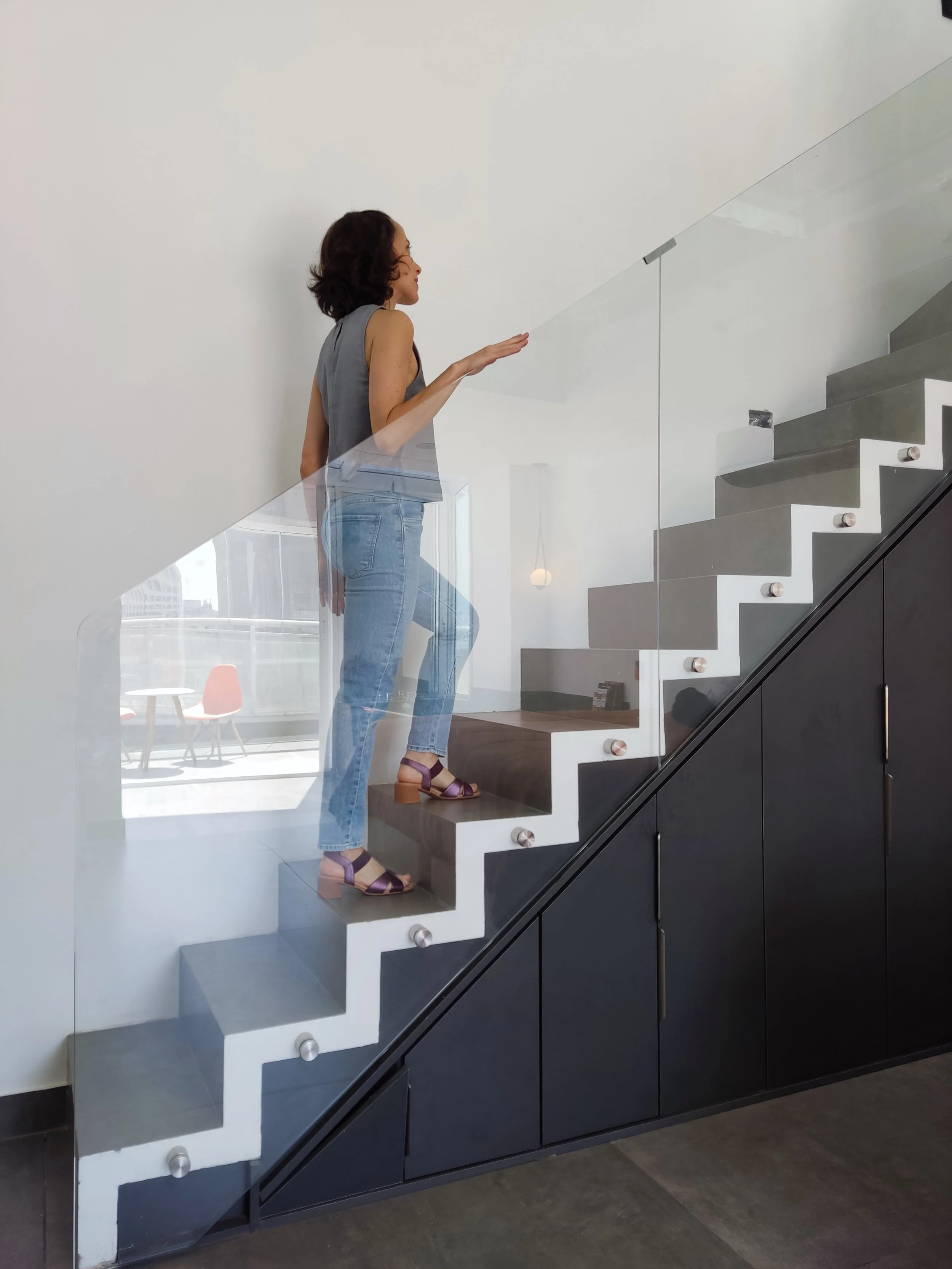 Mujer con cabello rizado y ropa casual en una escalera moderna con barandal de vidrio en interiores