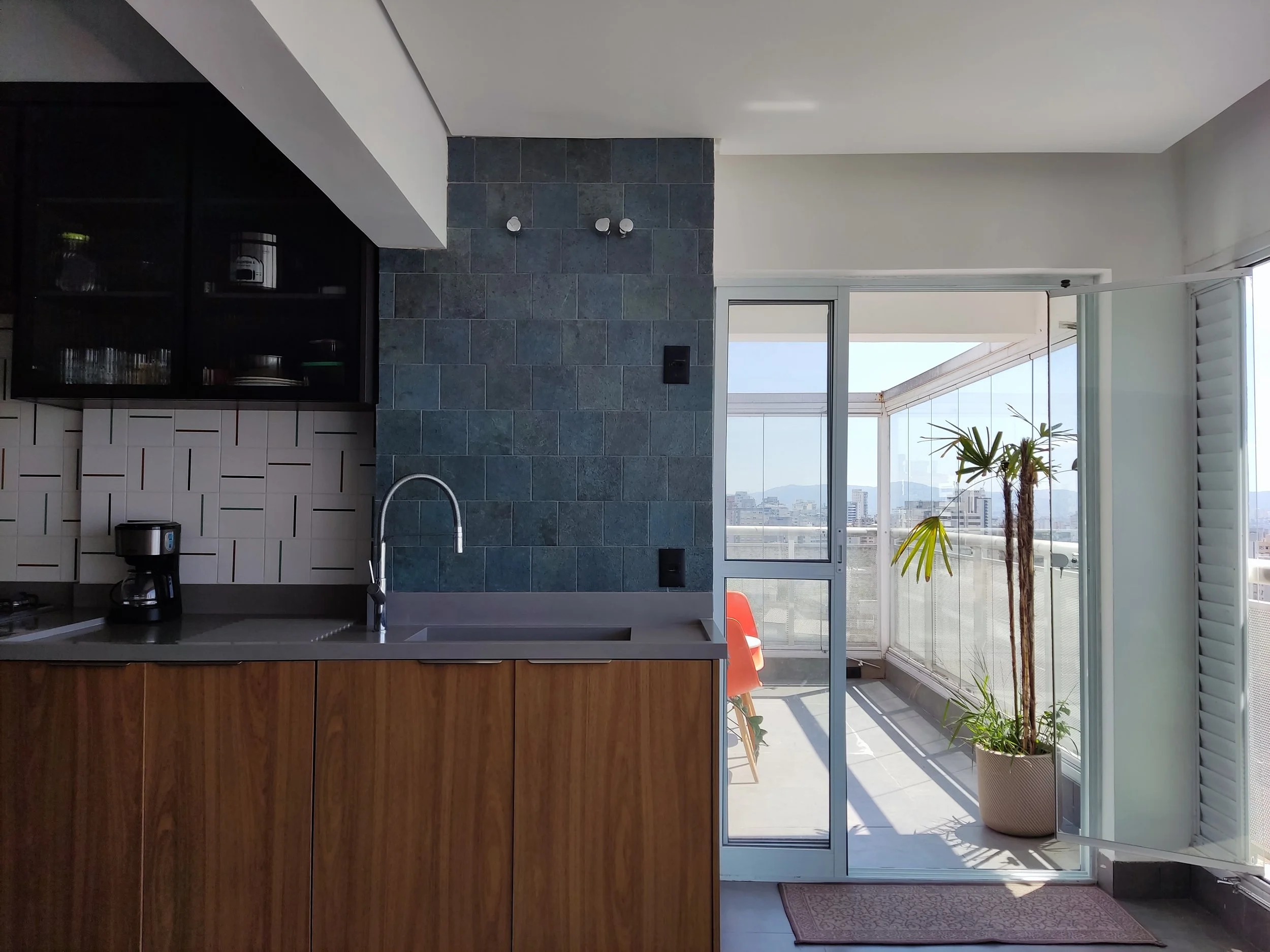 Cozinha moderna com bancada de madeira, parede de azulejos brancos e uma parede de tijolos de pedra cinza. Ao lado, uma porta de vidro que leva para uma varanda com plantas e uma vista da cidade.