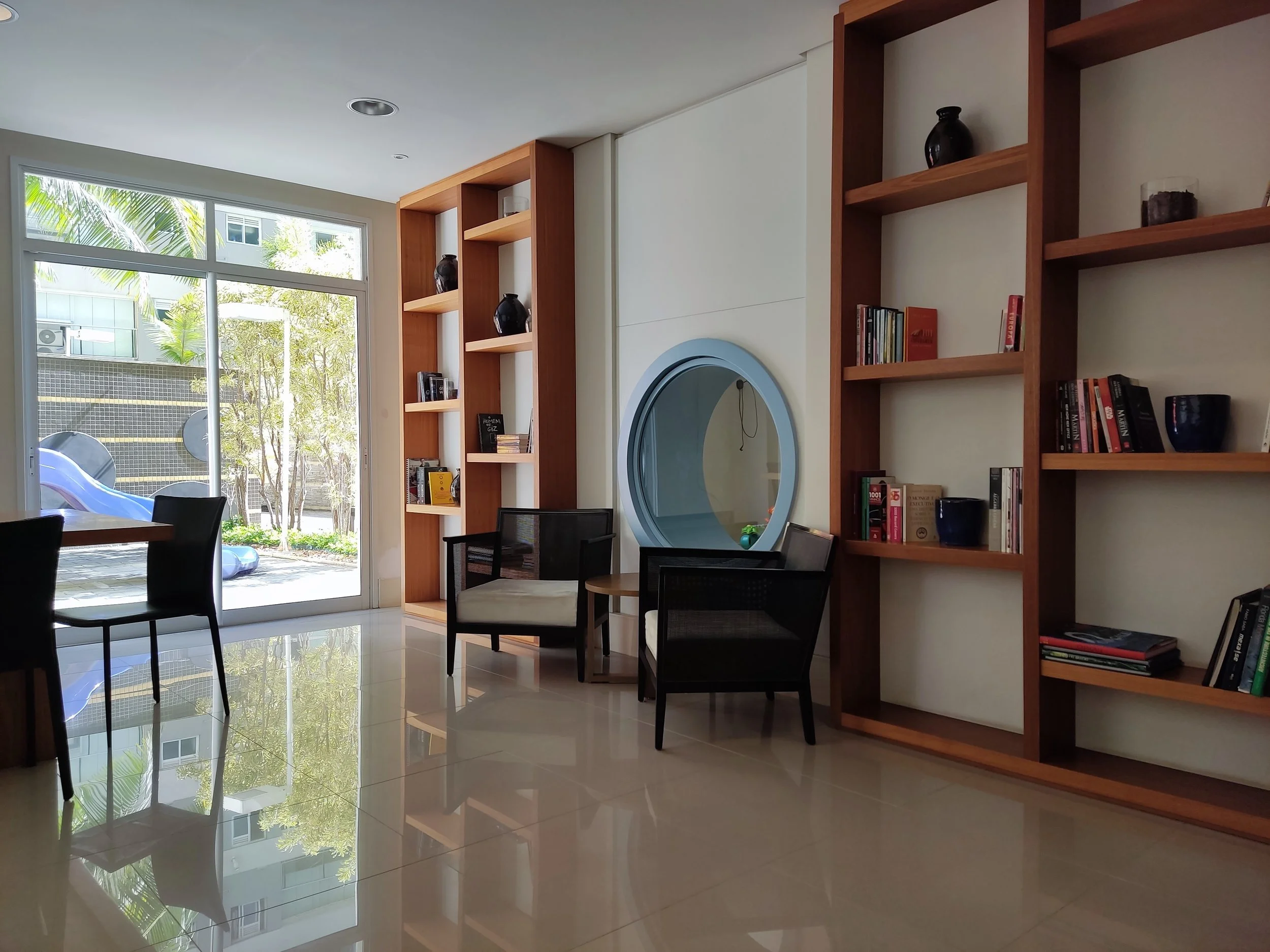 Sala com janelas grandes, móveis de madeira e cadeiras pretas, uma parede com estantes de madeira e uma janela redonda decorativa. Varios livros e vasos decorativos nas prateleiras, piso de azulejo brilhante.