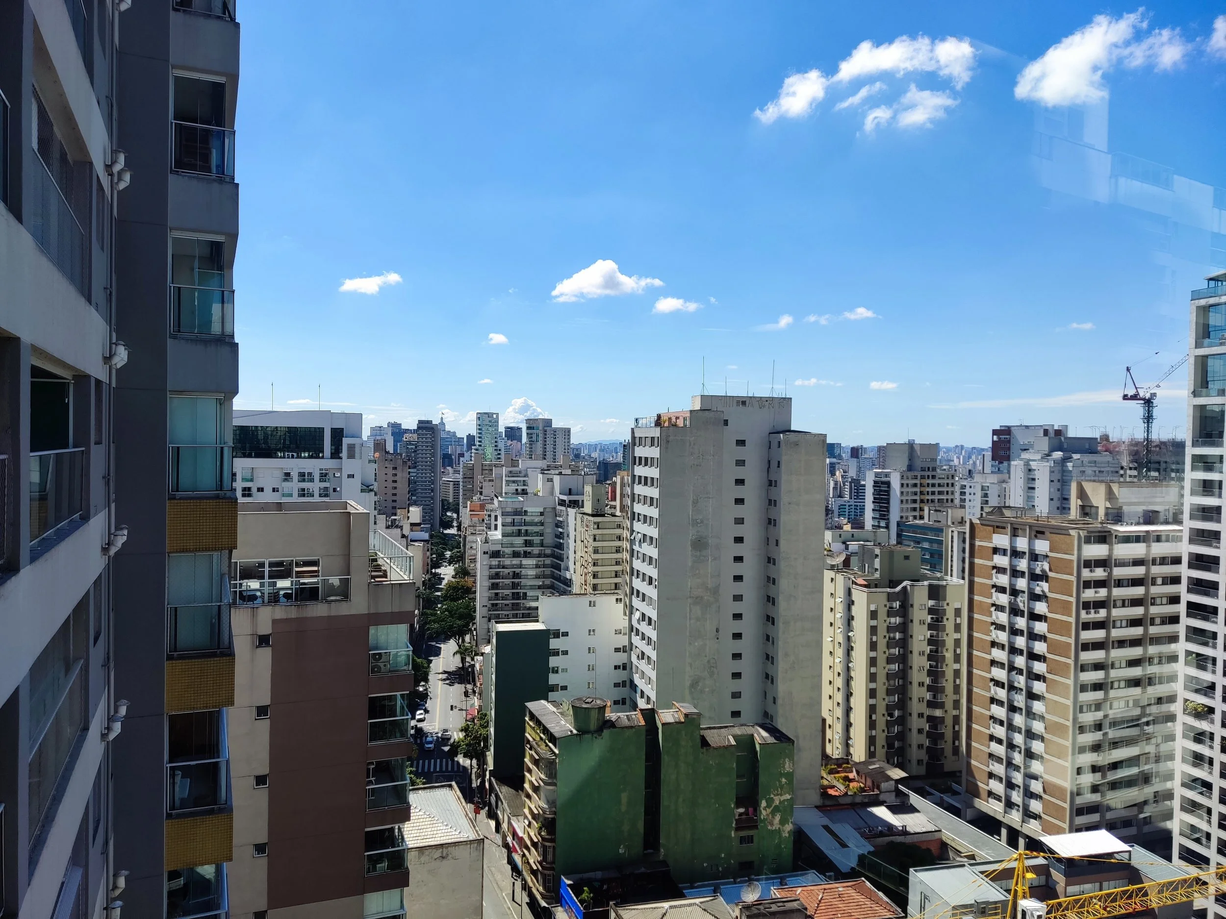 Vista de cidade com vários edifícios altos sob céu claro com algumas nuvens, incluindo prédios residenciais e comerciais de diferentes cores e tamanhos.