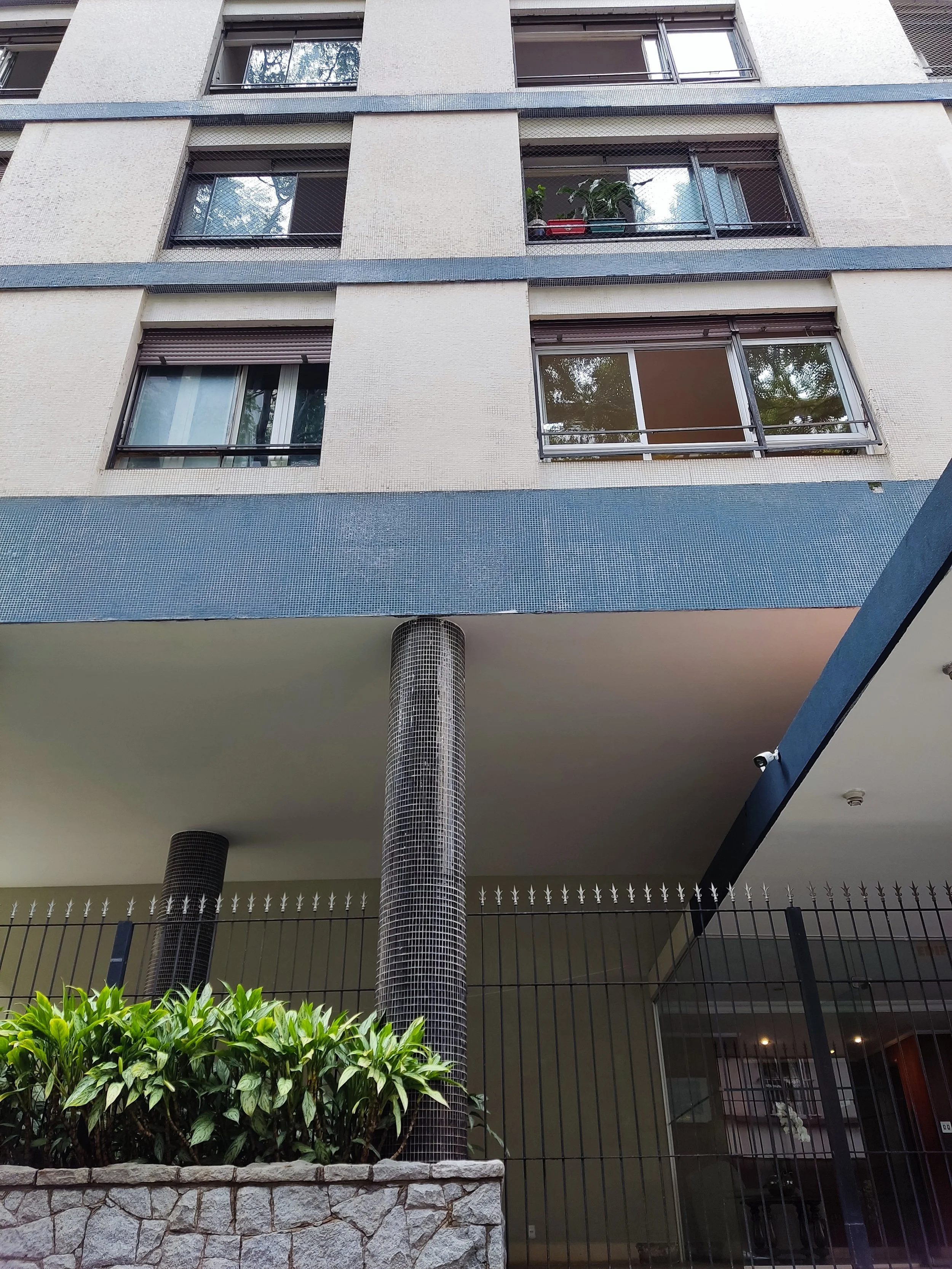 Foto da fachada de um prédio residencial com janelas, uma área de lazer, uma cerca preta, plantas verdes e colunas revestidas de mosaico.