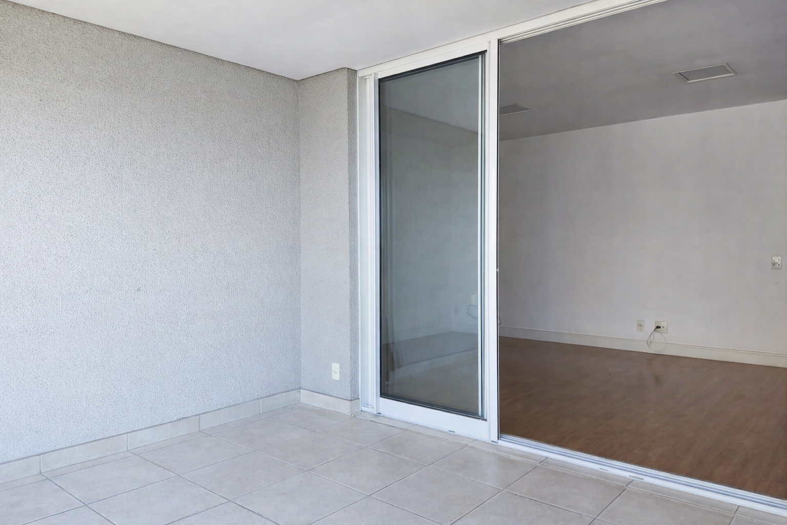 Varanda com piso de cerâmica e parede texturizada, com porta de vidro que dá acesso ao interior de uma sala com piso de madeira.
