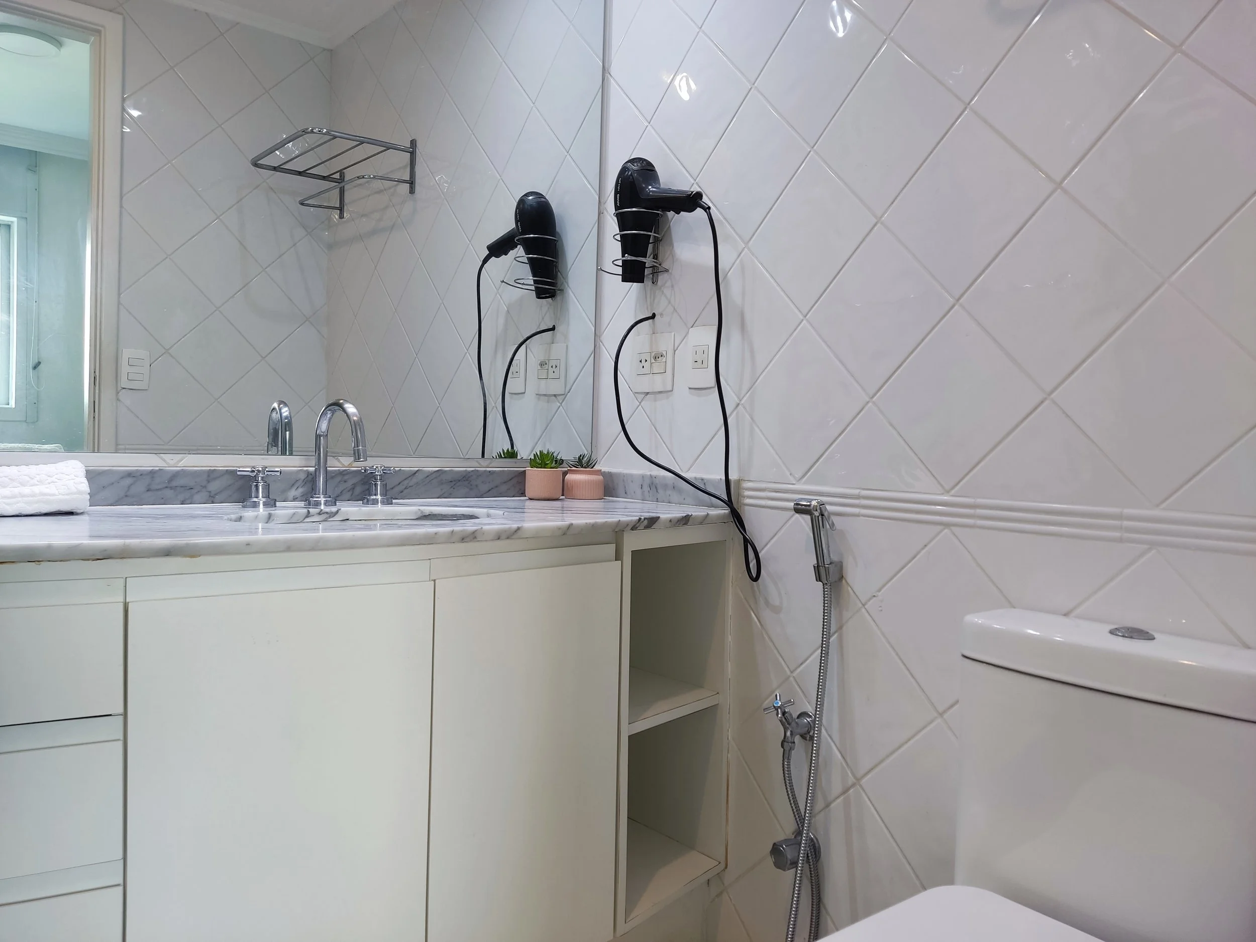 Banheiro com pia de mármore, espelho grande, cabelo no secador e pente, vaso sanitário, parede de azulejos brancos e uma janela ao fundo.