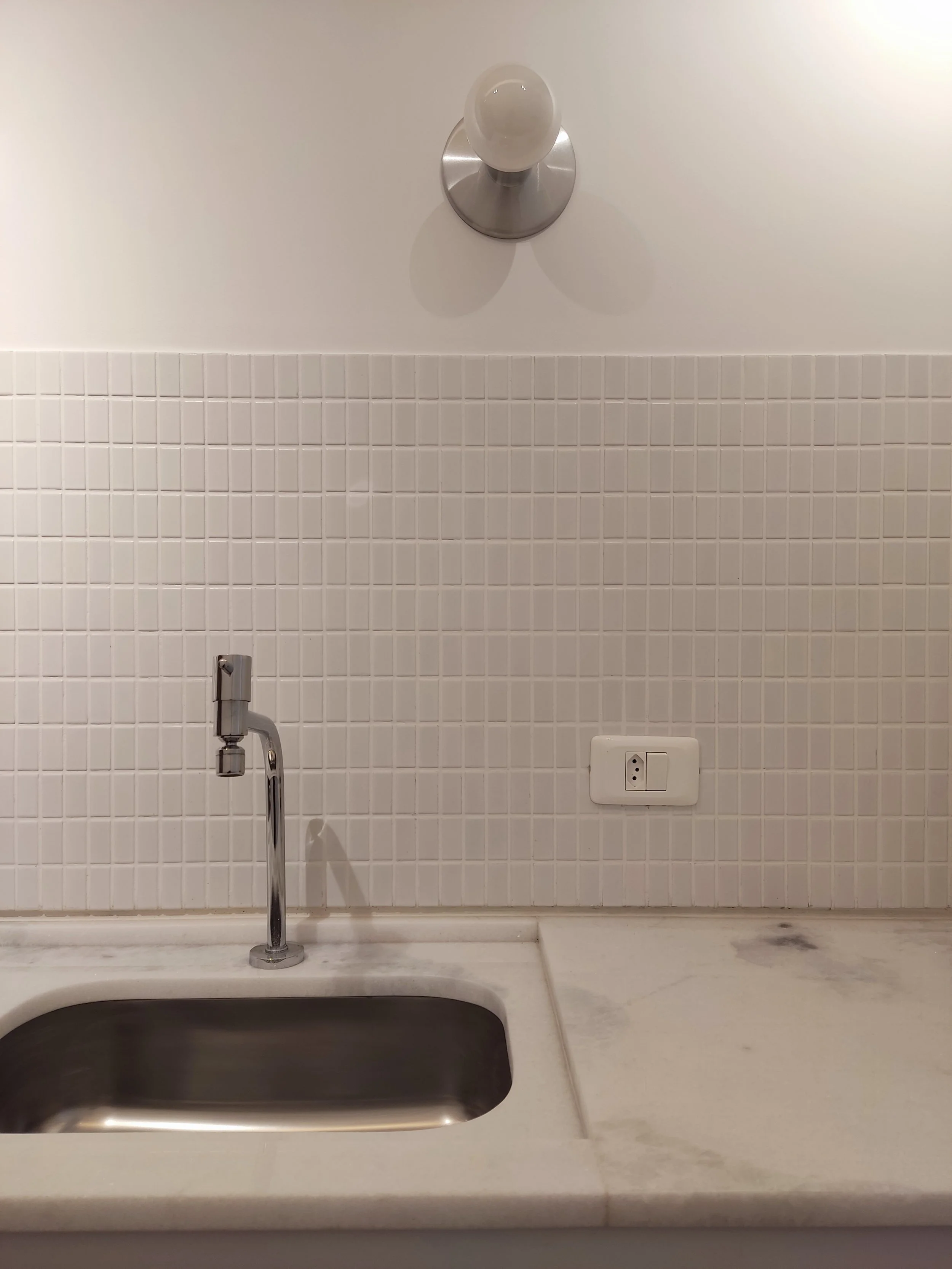 Pia de cozinha com pia de metal, torneira, parede com azulejos brancos e tomada elétrica.