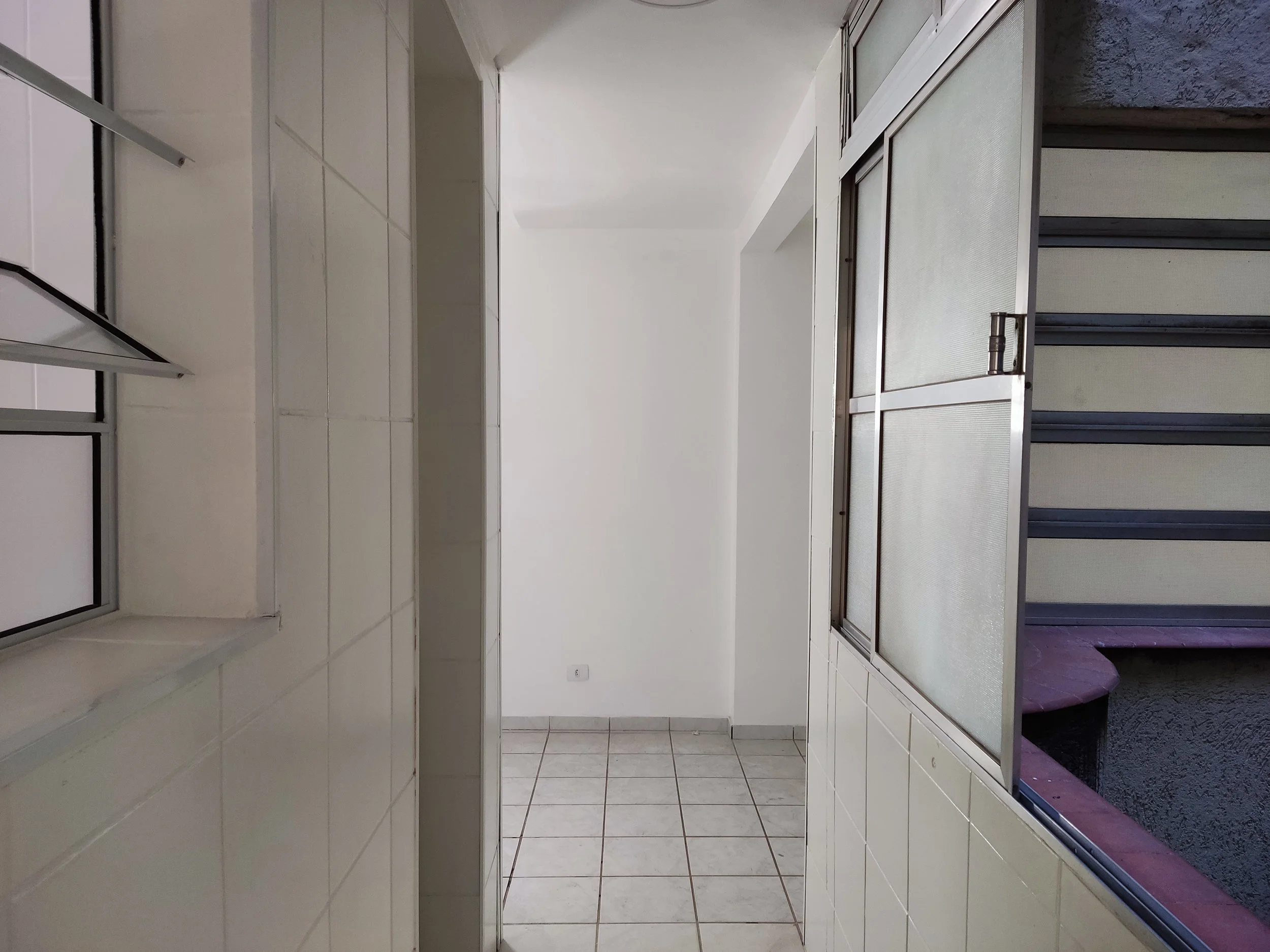 Corredor de apartamento com paredes brancas, piso de azulejo branco, janela de vidro com moldura de alumínio e uma varanda ou espaço externo visível ao lado.