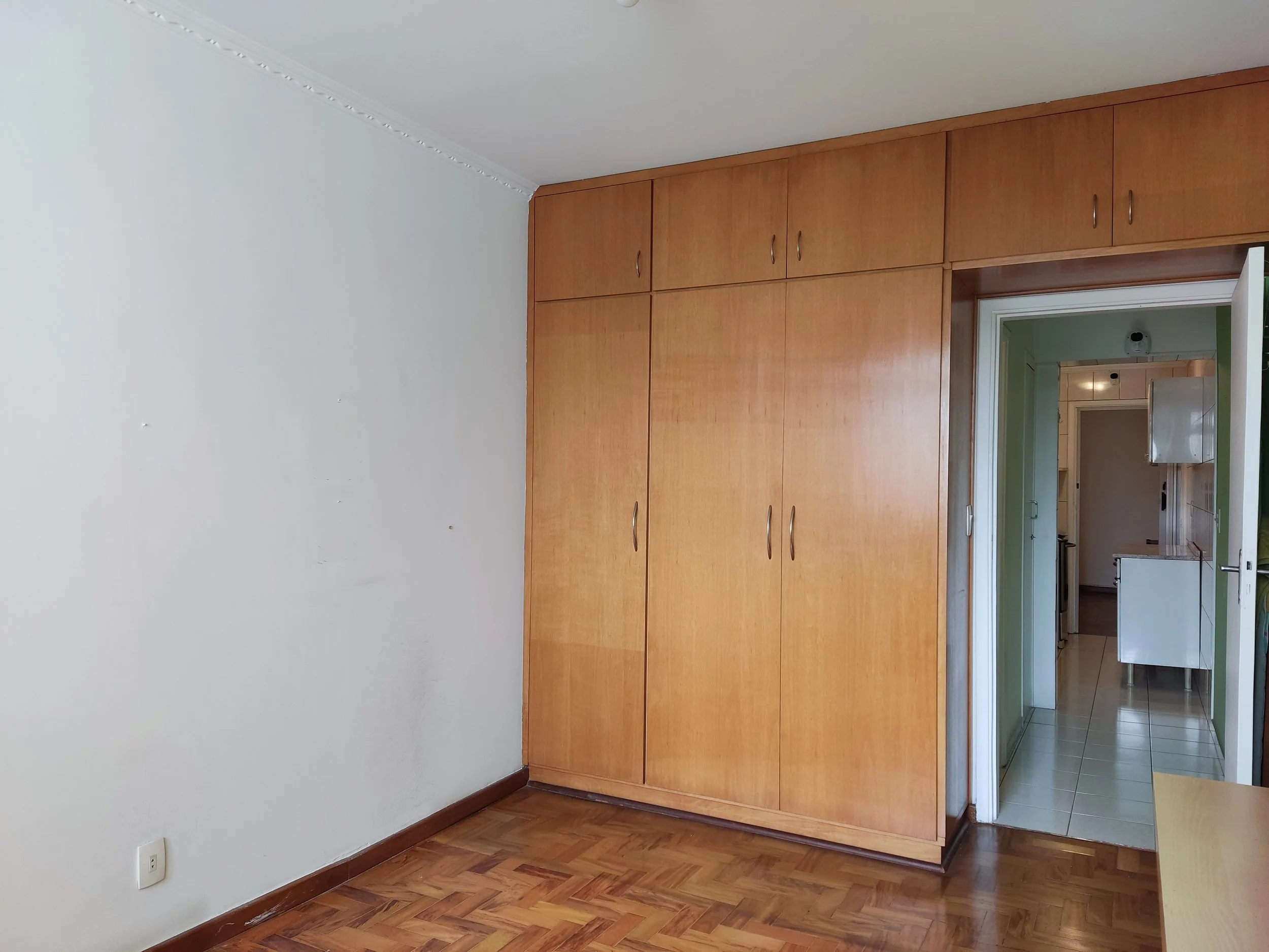 Quarto sem móveis com parede branca, piso de madeira e armário de madeira ao lado da porta que leva à cozinha, que tem piso de azulejo e armários brancos.