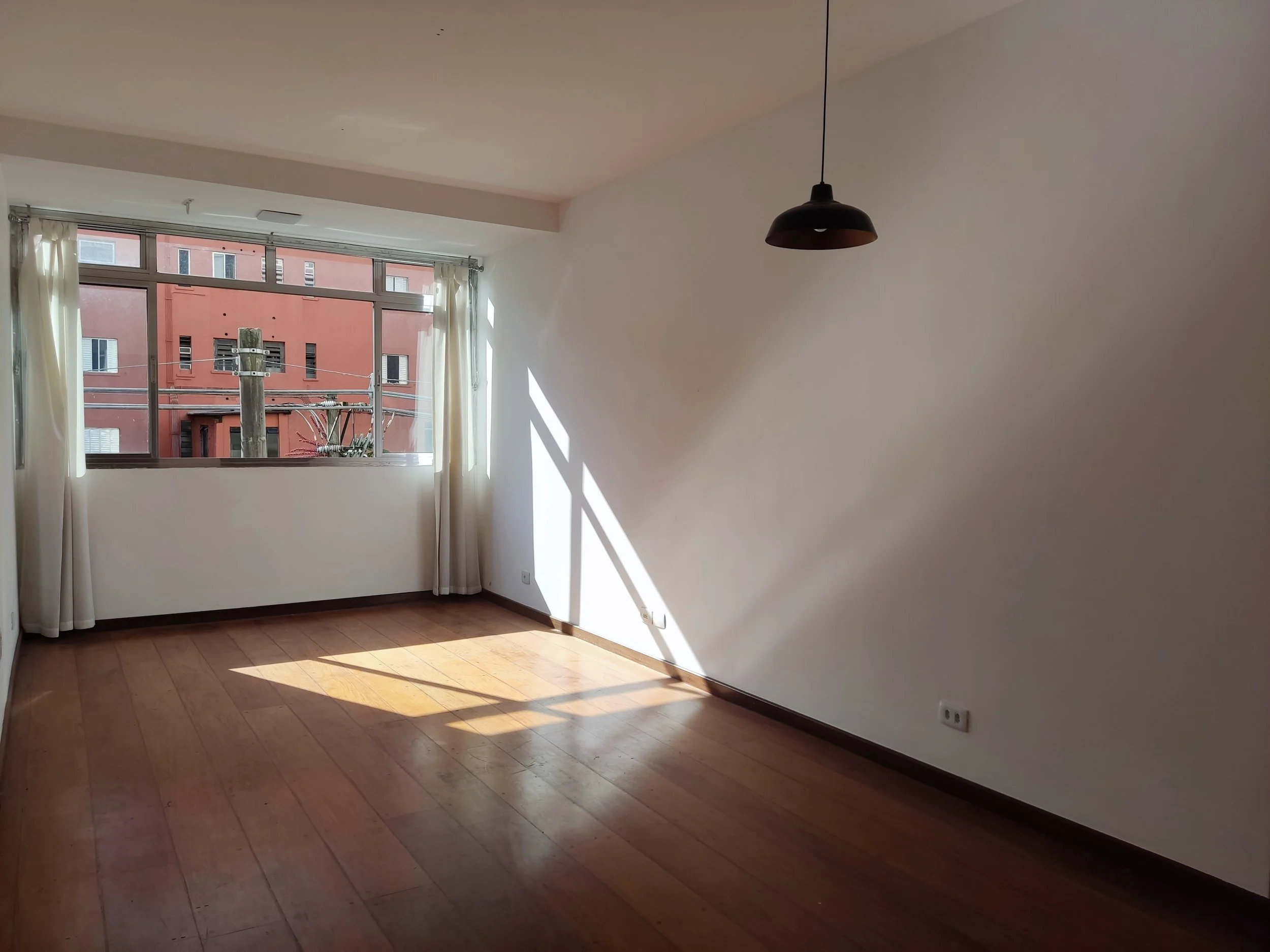 Sala vazia com piso de madeira, parede branca, grande janela com cortinas claras e luminária de teto preta.