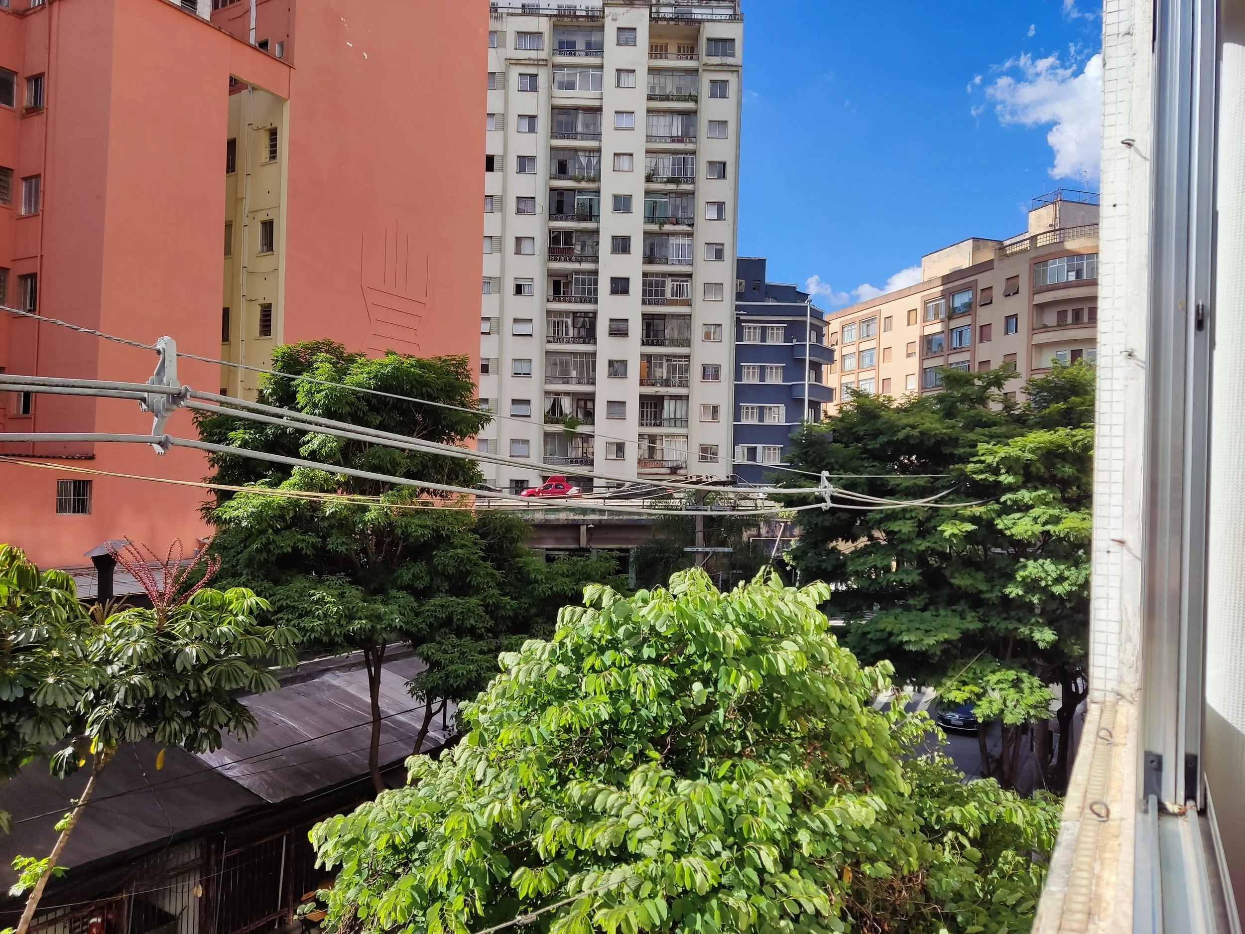 Vista de prédios altos de cor laranja, azul e branco ao fundo, com árvores verdes na frente e fios de varal suspensos entre os prédios, sob céu azul com algumas nuvens brancas.