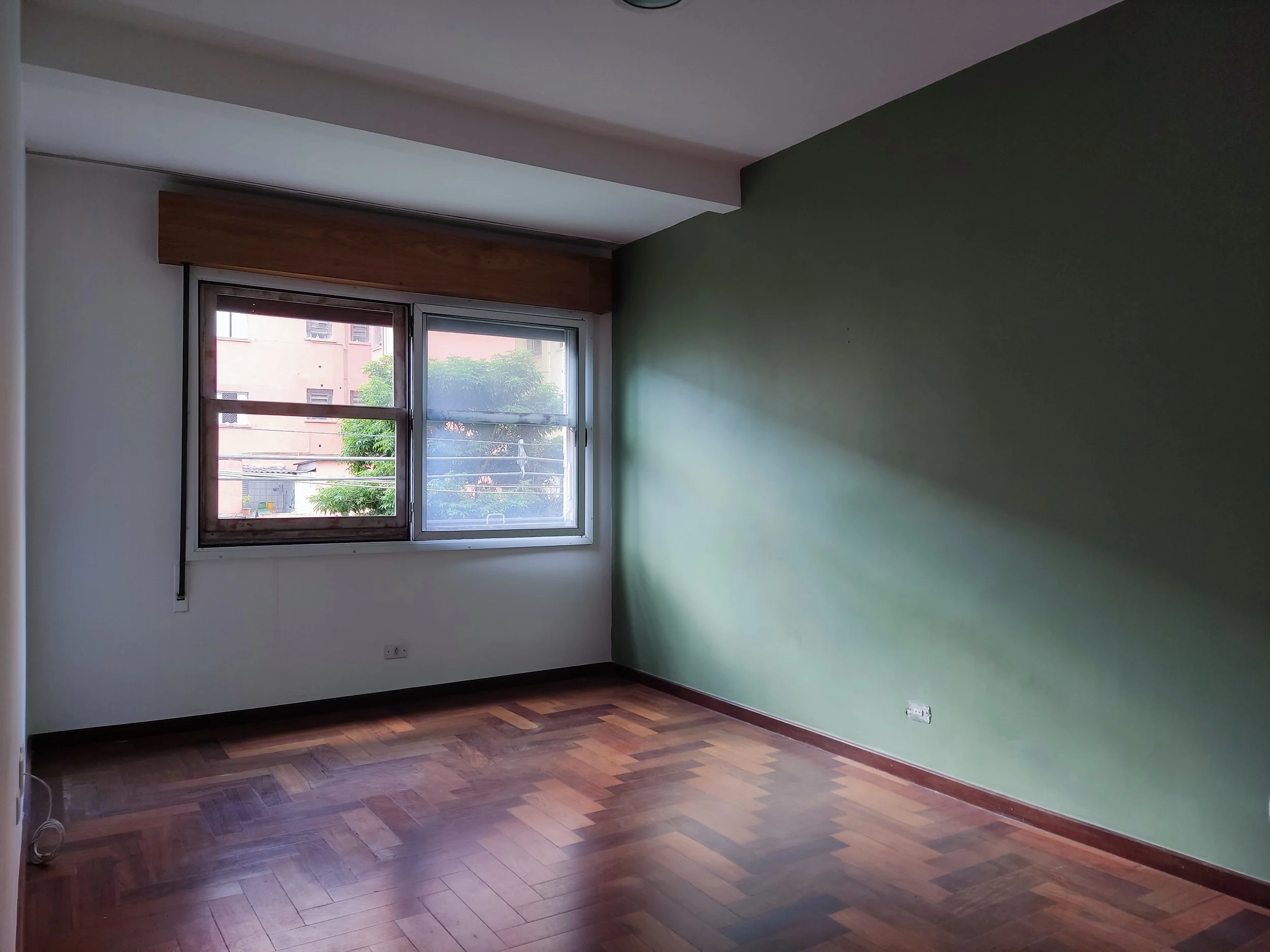 Quarto vazio com janela grande, parede verde e piso de madeira.