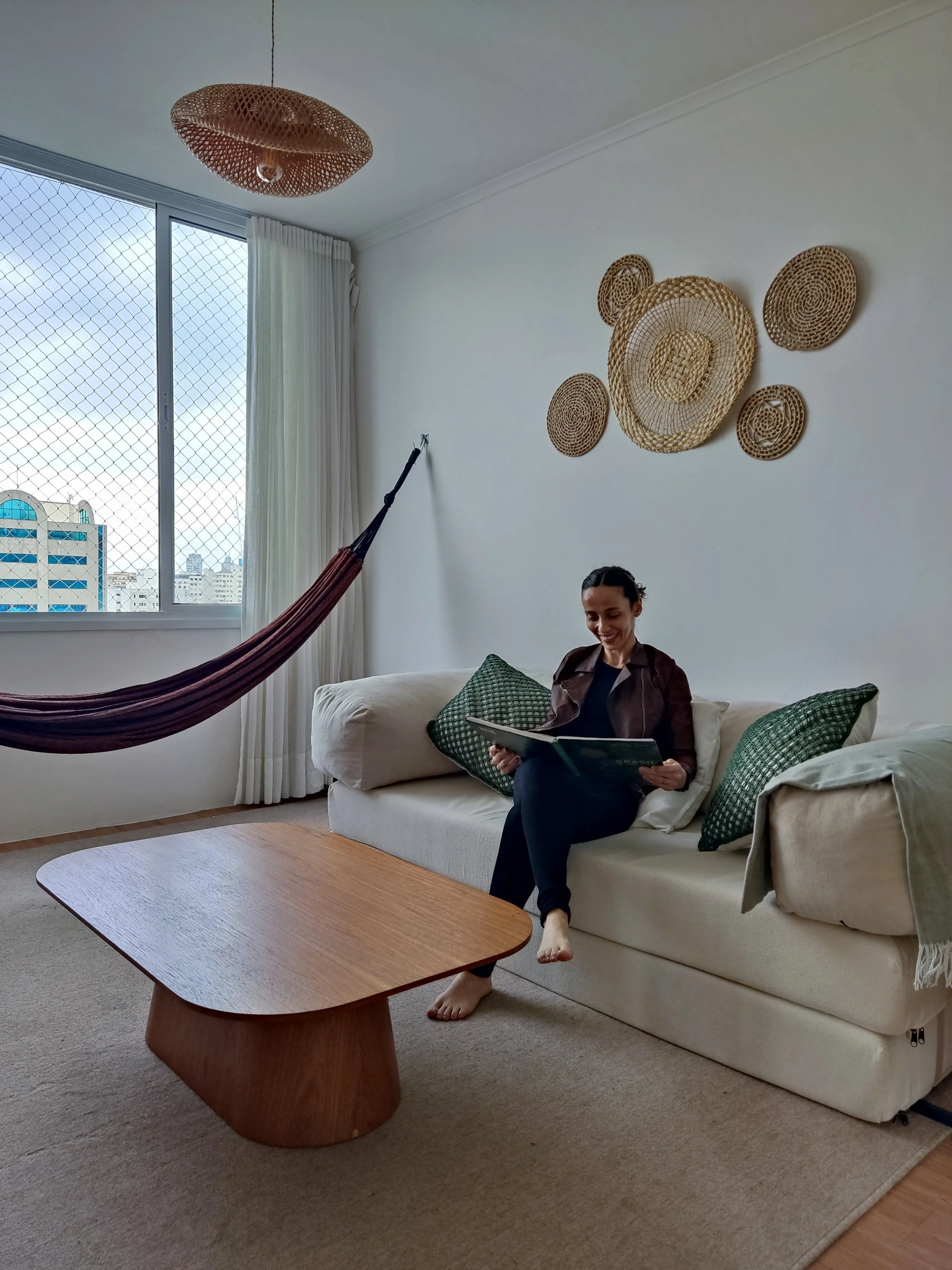 Mulher sentada no sofá de casa, lendo um livro ou revista, com almofadas verdes ao redor, em um ambiente decorado com cestos de palha na parede, uma rede de descanso e uma janela com vista para prédios.