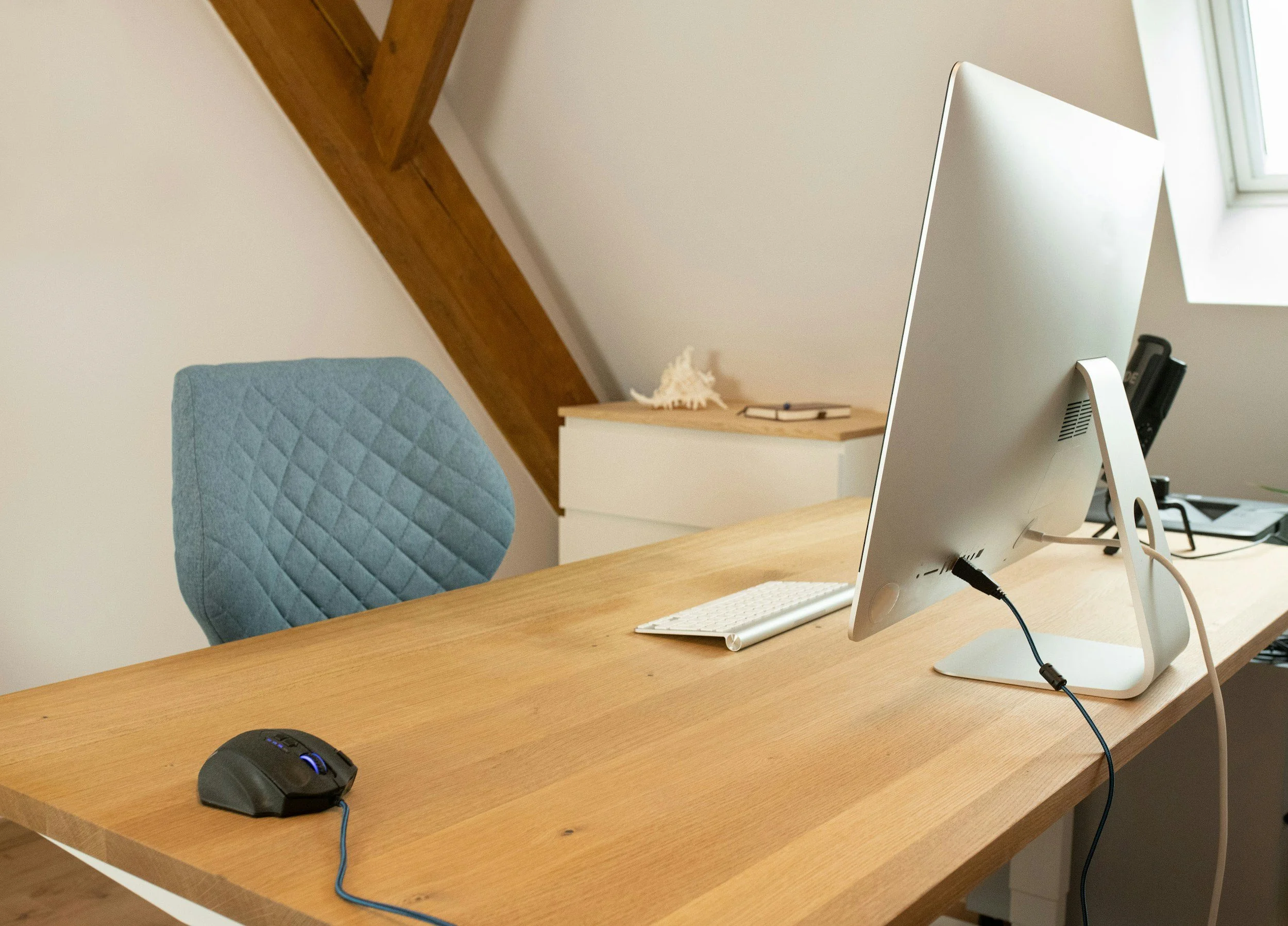 Home office desk with a computer monitor, keyboard, mouse, and a blue cushioned chair in a room with sloped ceiling and a window.