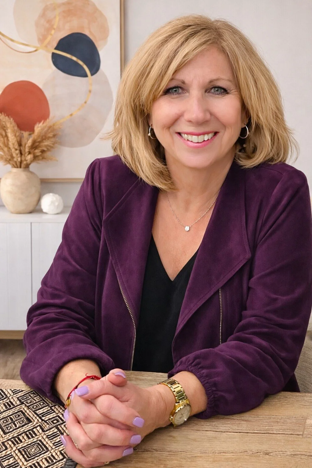 A woman with blonde hair, wearing a purple jacket, smiling, sitting at a table.