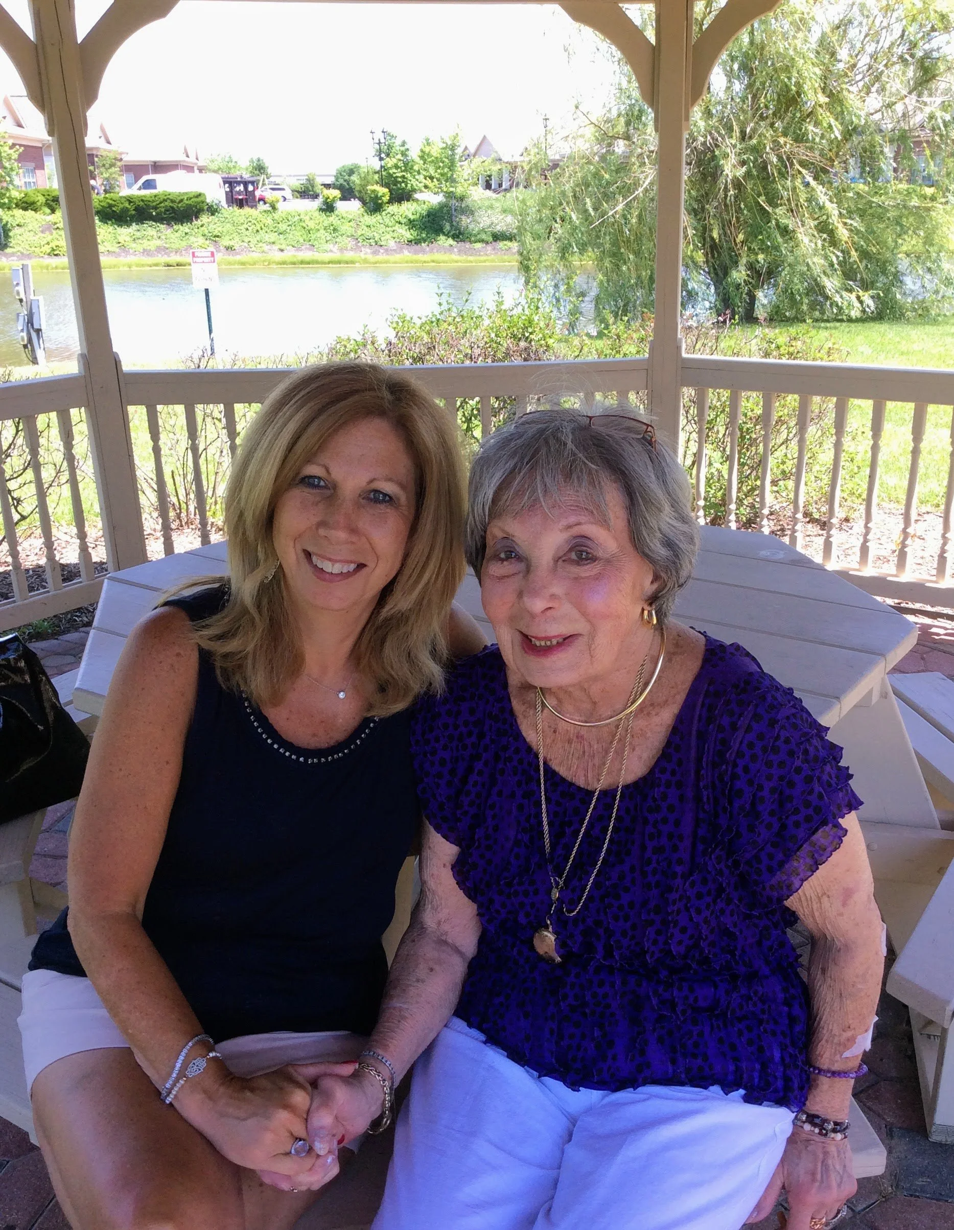 Two women sitting together on a park pavilion bench, holding hands, with a pond and trees in the background.
